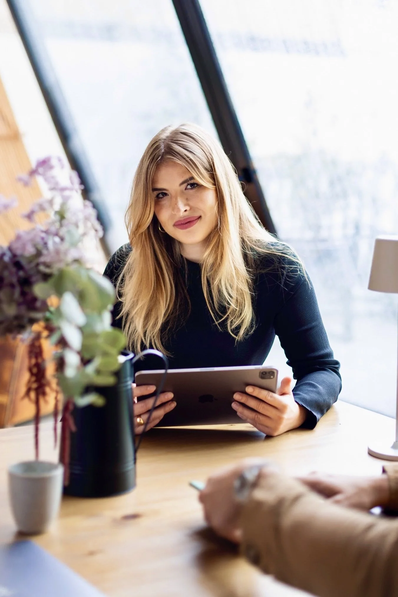 Une femme blonde tenant une tablette, assise à une table en bois avec des fleurs et une lampe à côté, dans une pièce lumineuse.