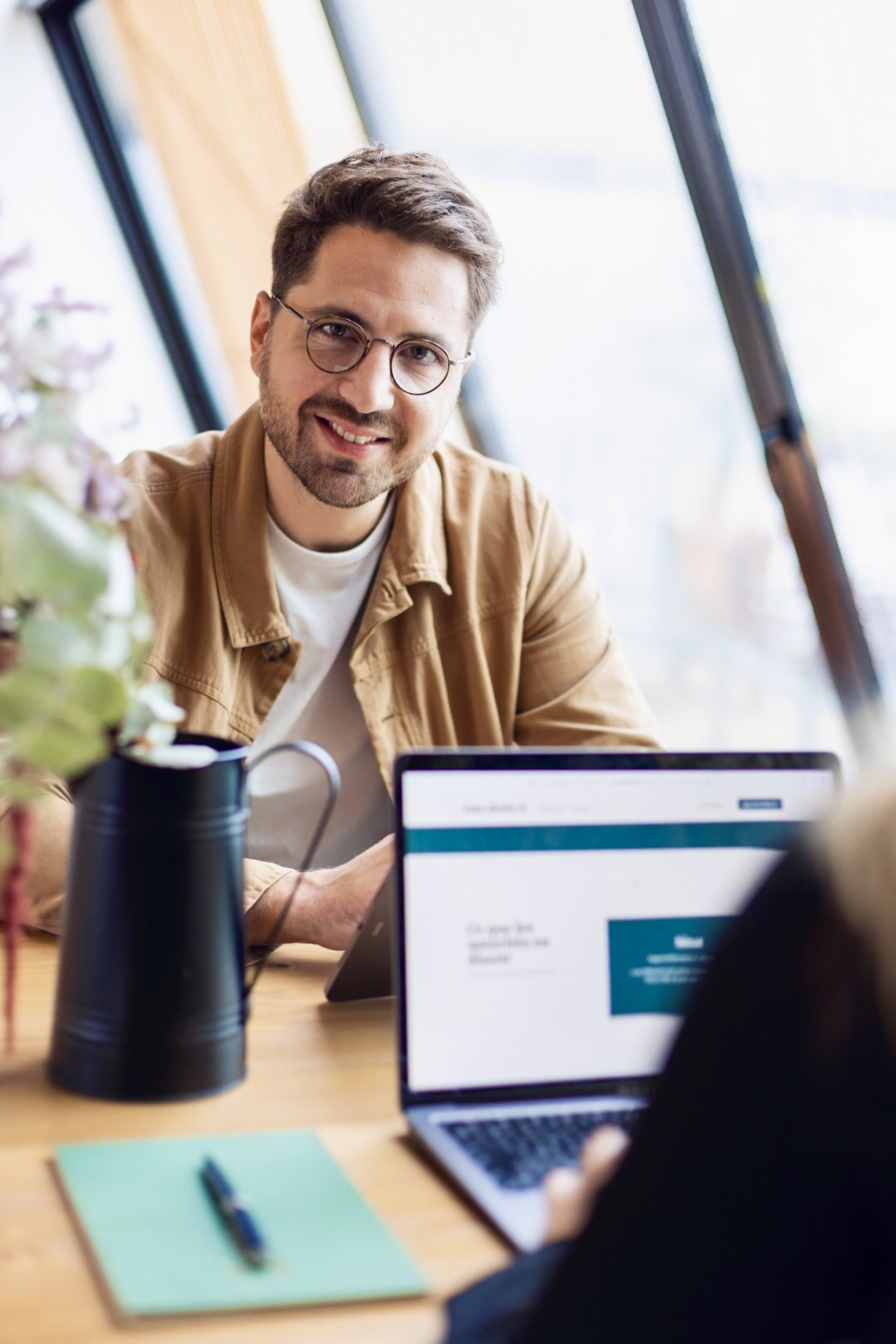 Jeune homme souriant, portant des lunettes, assis à une table, face à une personne utilisant un ordinateur portable, dans un environnement lumineux avec de grandes fenêtres.