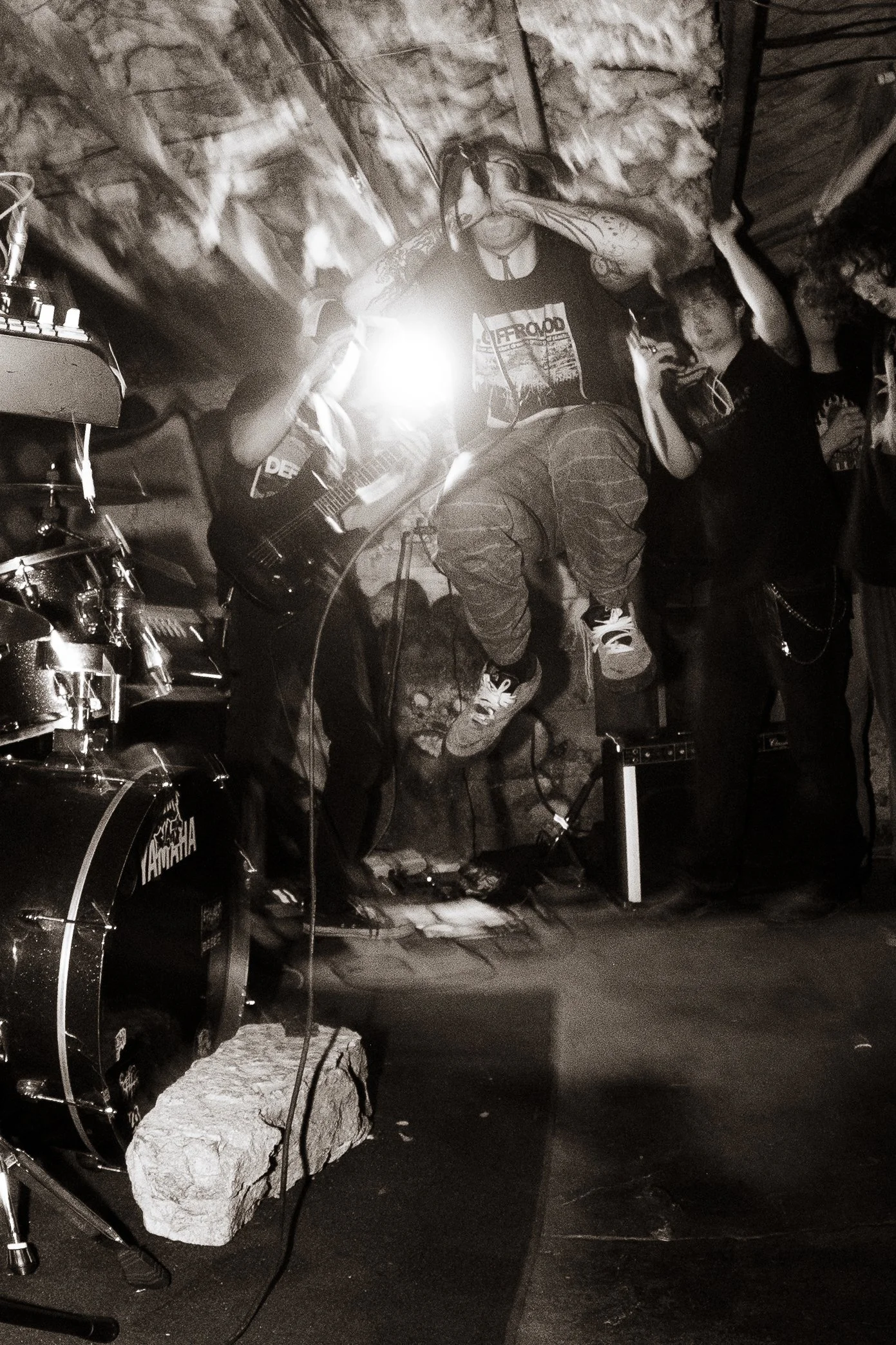 A band performing on stage with a drummer and guitarist, with one singer jumping and singing into a microphone. The scene is dark with a stone wall background, and the image is in black and white.
