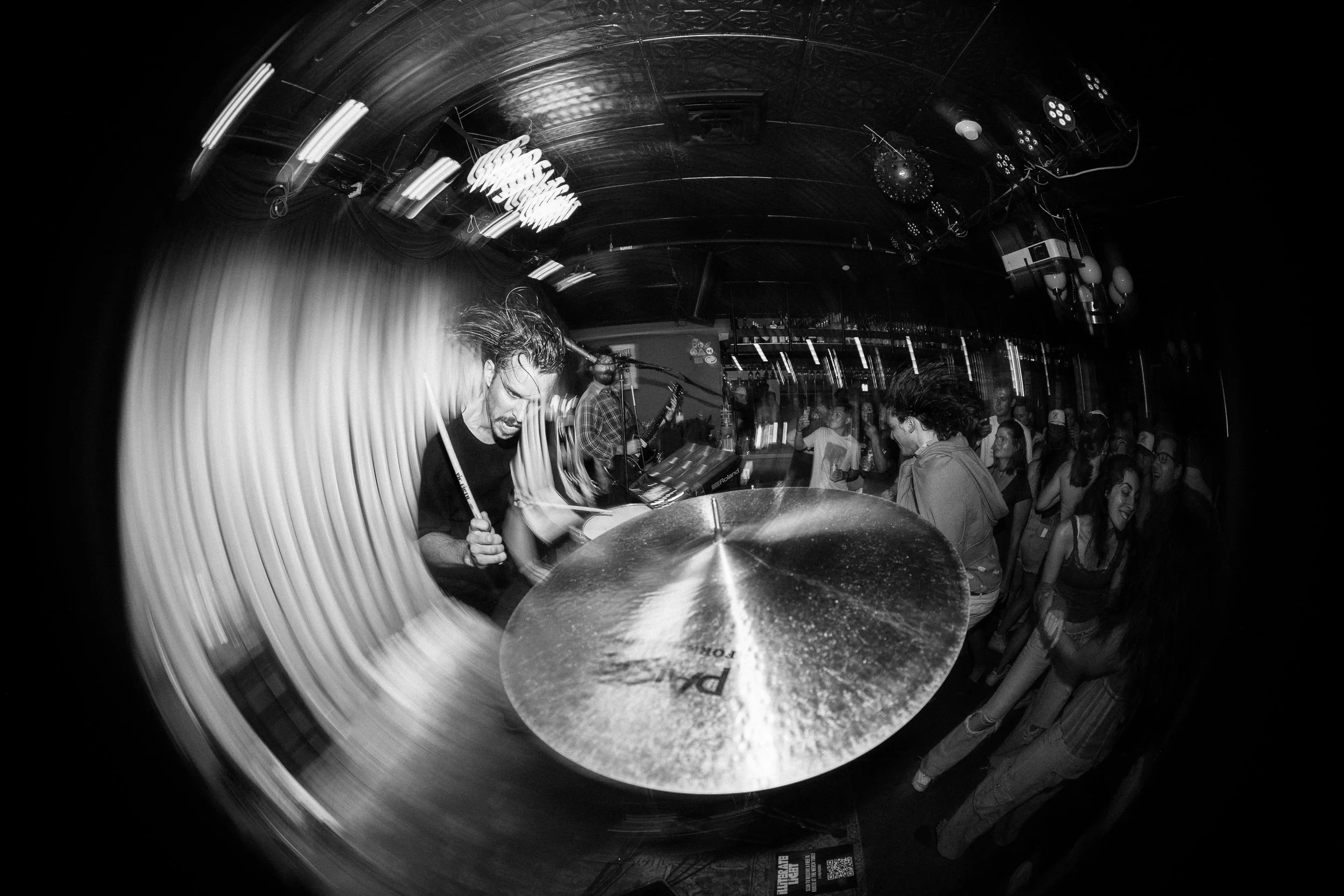 Black and white photo of a live music performance in a venue with a crowd of people dancing and enjoying the show, with a focus on a drummer in the foreground and a guitarist in the background.