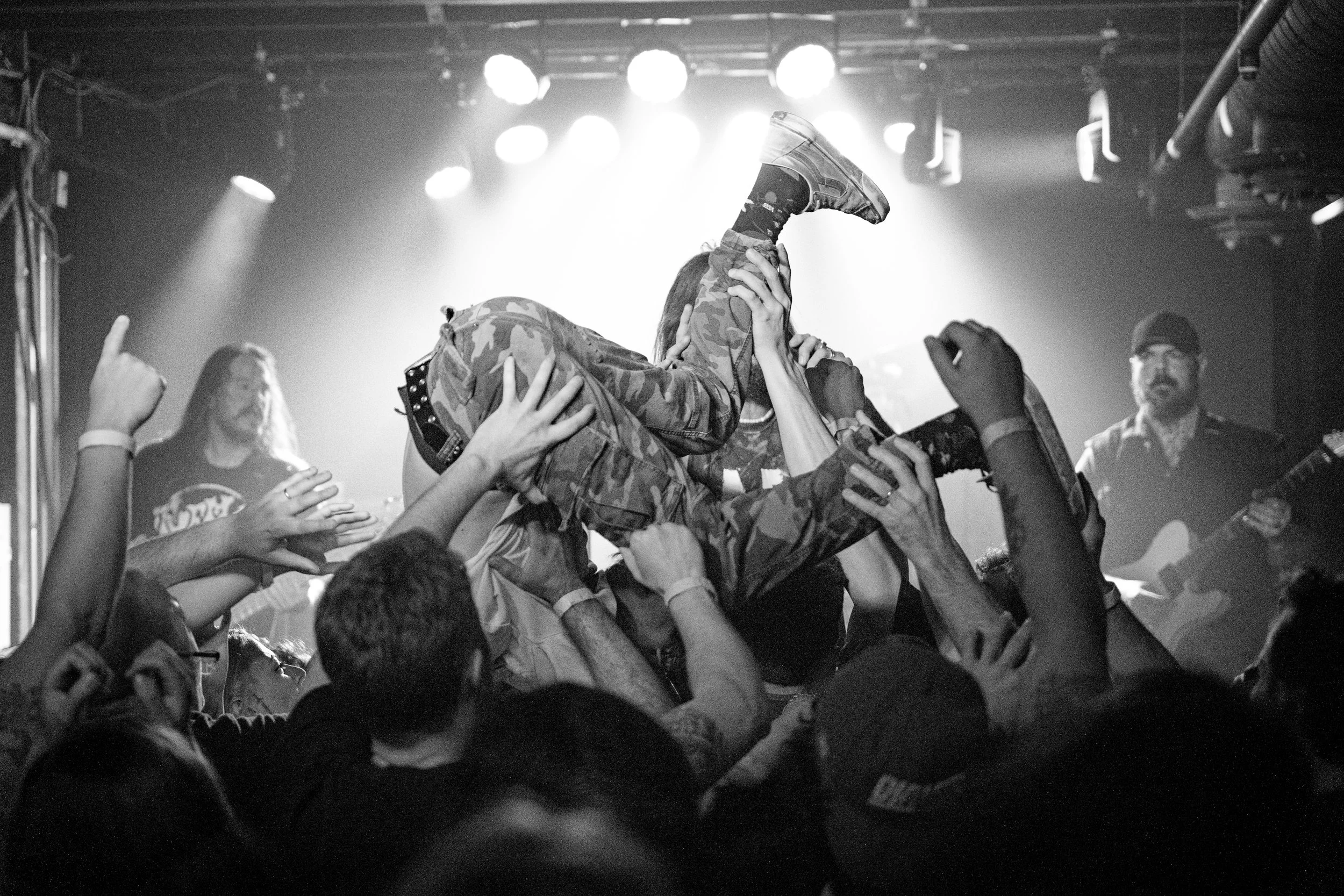 A black and white photo of a concert scene with a crowd lifting a person. The performer is wearing camouflage pants and sneakers, and the audience is reaching out to support them. There are stage lights and two musicians in the background playing gui