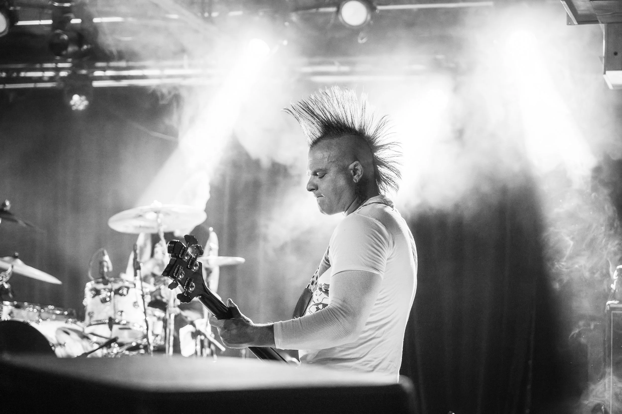 A punk rock musician with spiky hair playing a bass guitar on stage during a concert, with bright stage lights and a drum set in the background.