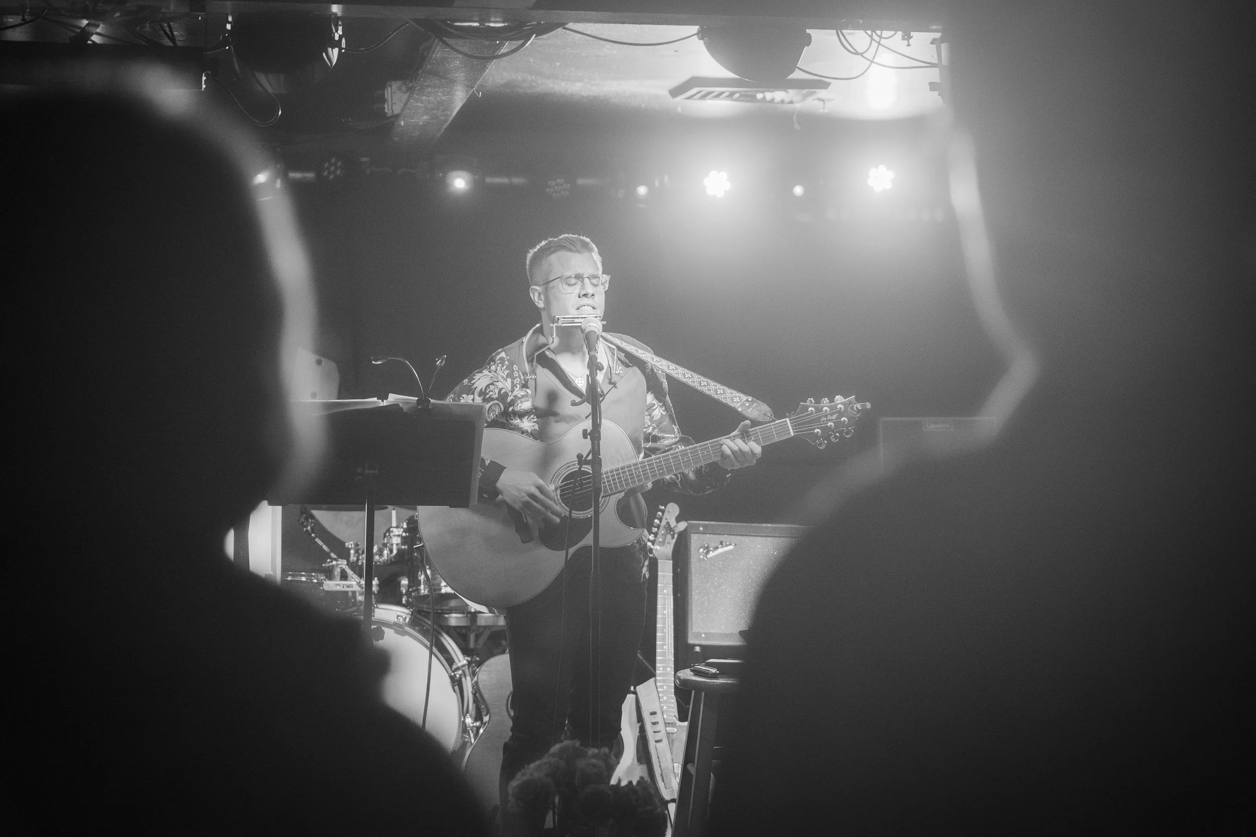Black and white photo of a male musician performing on stage with an acoustic guitar and harmonica, in front of an audience, with stage lights overhead.