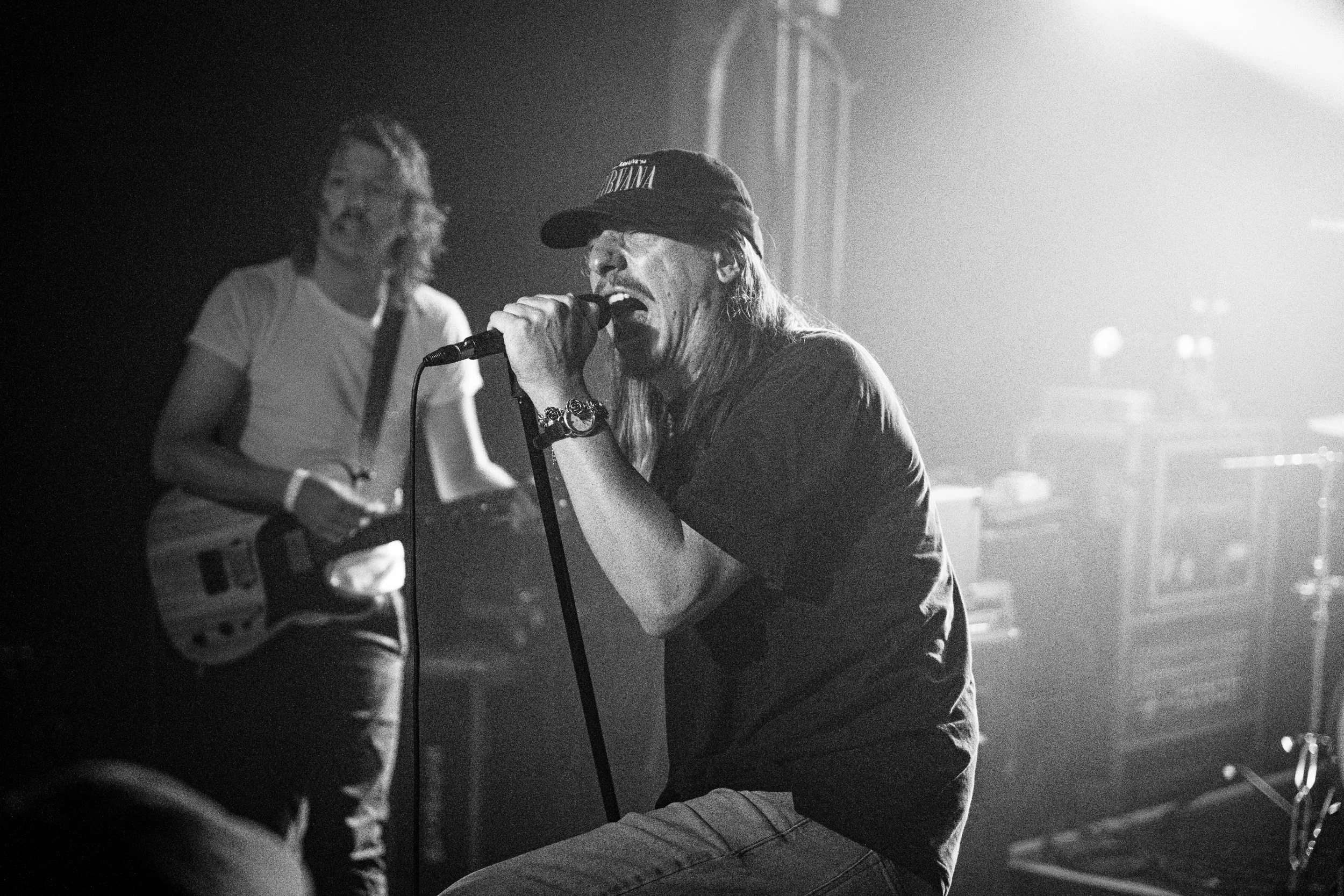 A black and white photo of a musician singing into a microphone during a live concert. The singer is wearing a cap and casual clothing, with long hair and a bracelet. In the background, another person, likely a guitarist, is seen playing the guitar, 