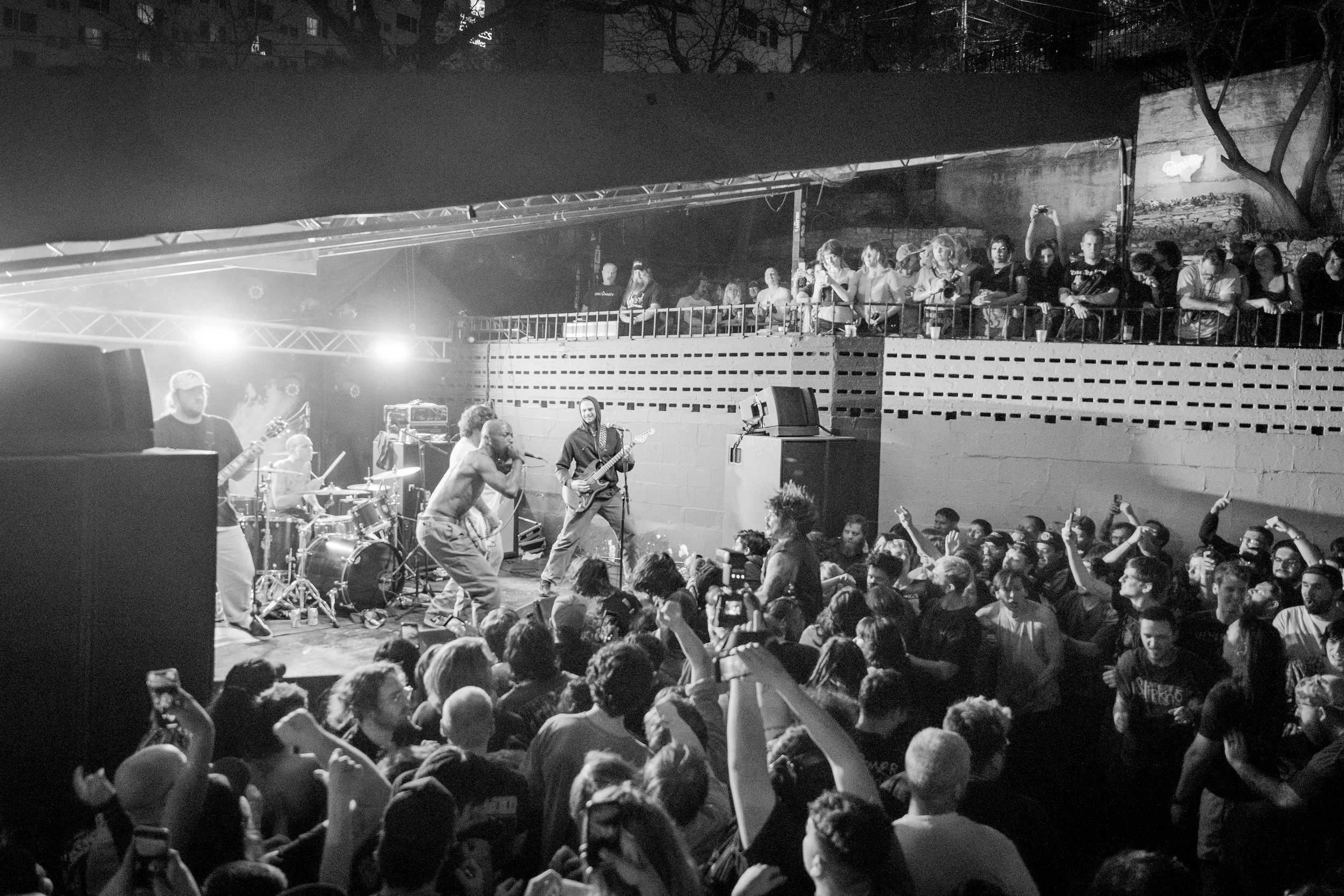 A black and white photo of a live outdoor concert showing a band performing on stage with an enthusiastic crowd, some raising their hands, and an audience seated on an elevated platform in the background.