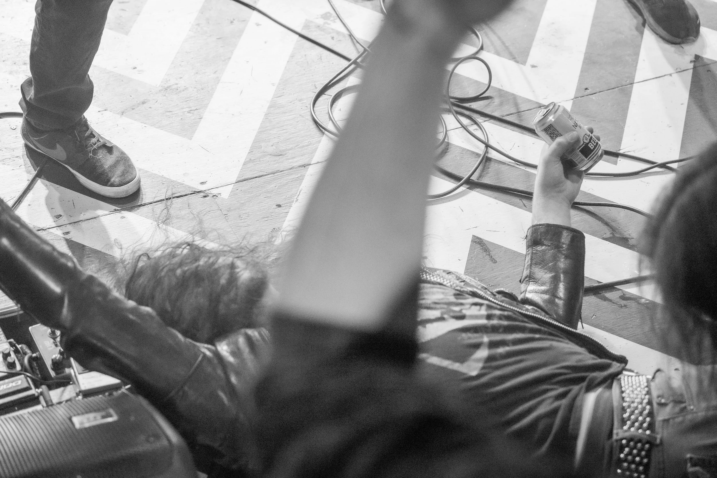 A person lying on the floor, holding a soda can, with some wires and a microphone nearby. The floor has painted arrow markings.