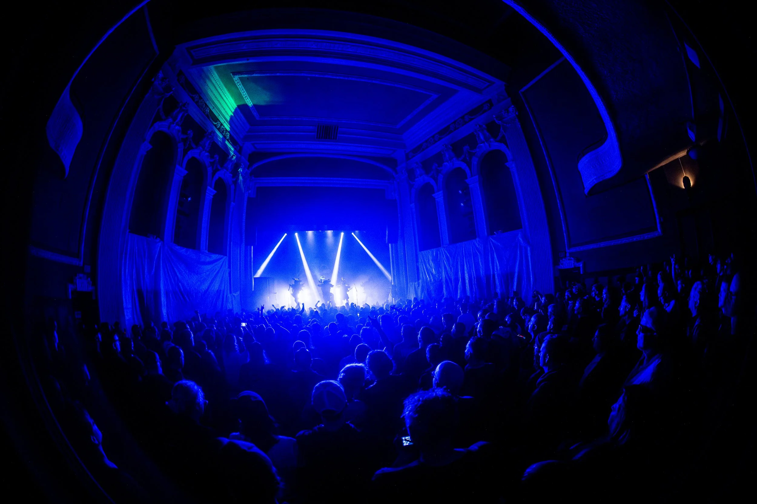 An indoor concert with a large audience, stage lights, and a band performing, in a historic theater with ornate interior details.