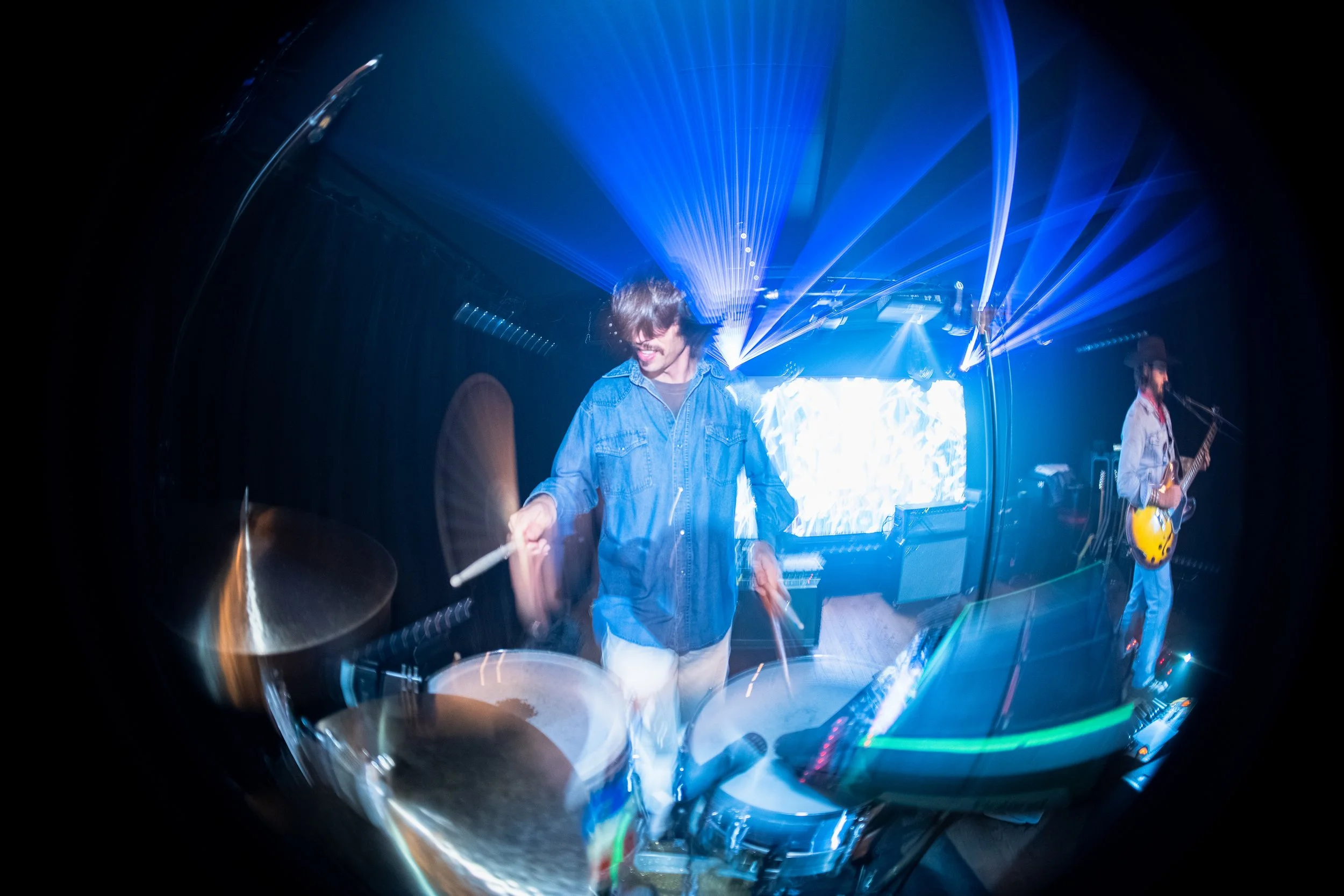 Musicians performing on stage with blue laser lights and abstract lighting effects, one playing drums and another with a guitar.