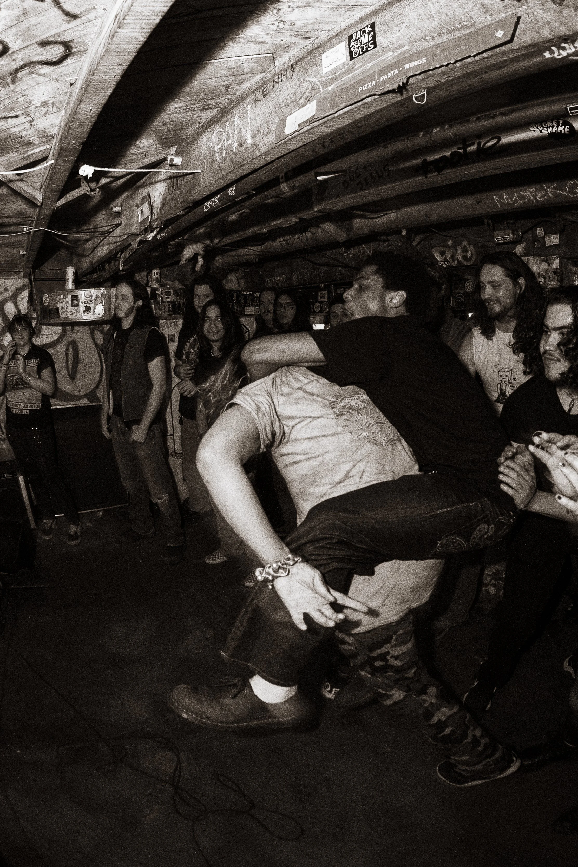 A group of people at a music venue enjoying a mosh pit, with one person crowd surfing, under dim, graffiti-covered ceiling lighting.