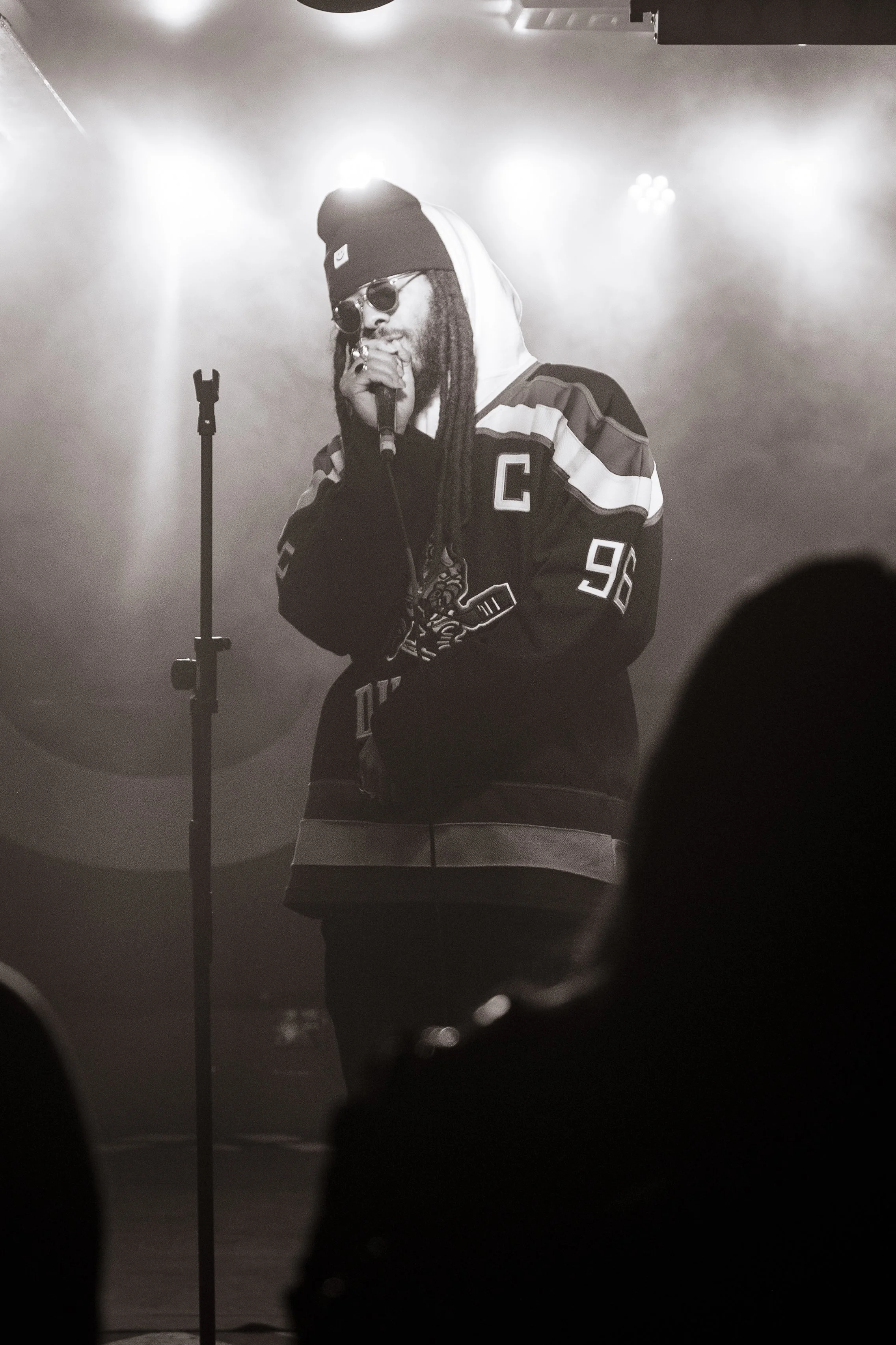 A musician with dreadlocks wearing sunglasses, a beanie, and a hockey jersey singing into a microphone on stage with bright lights and fog.