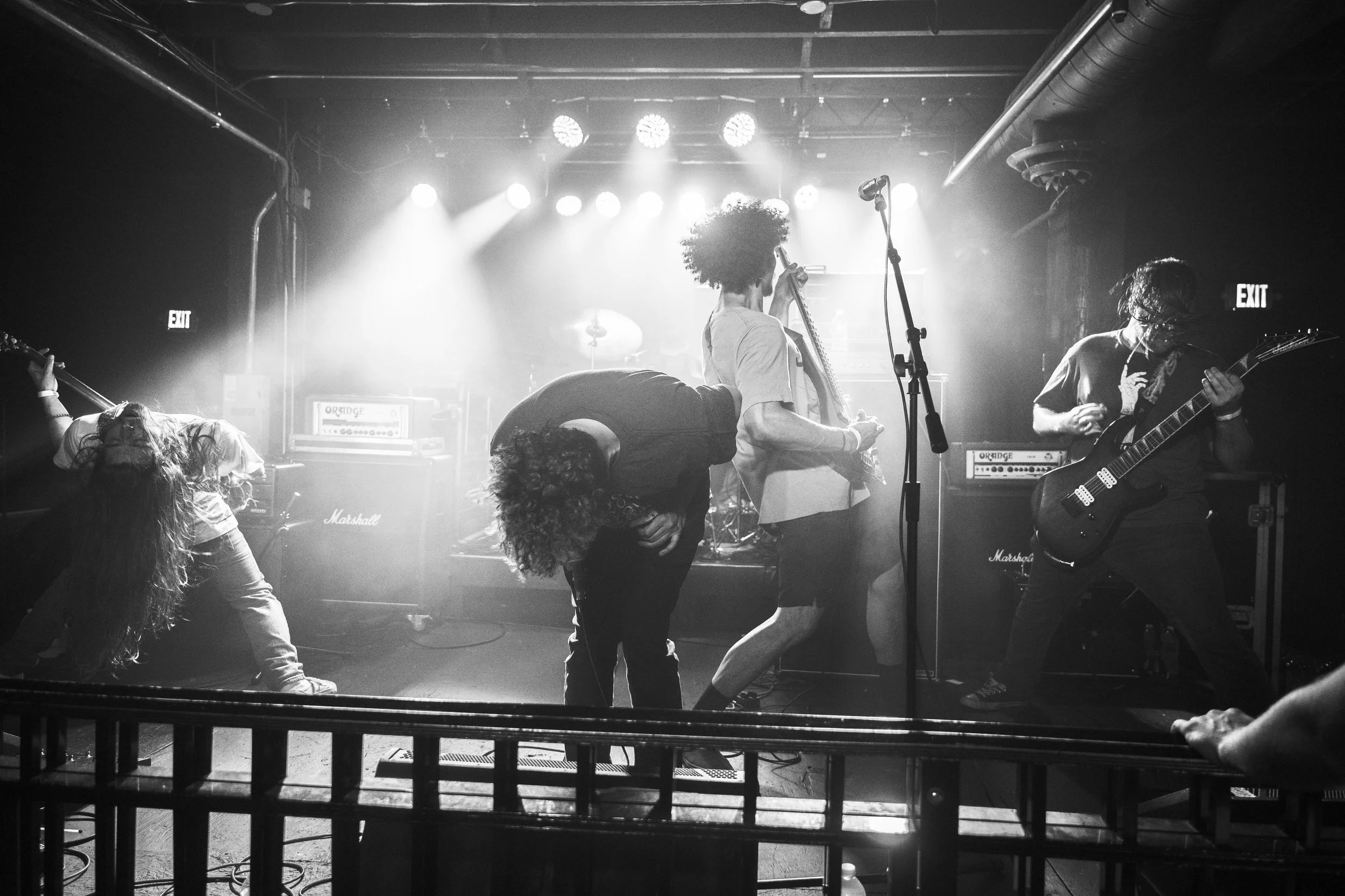 Black and white photo of a band performing on stage with three musicians. One is playing guitar, another singing into a microphone, and a third is bent over in the left background. Bright stage lights are shining behind them.