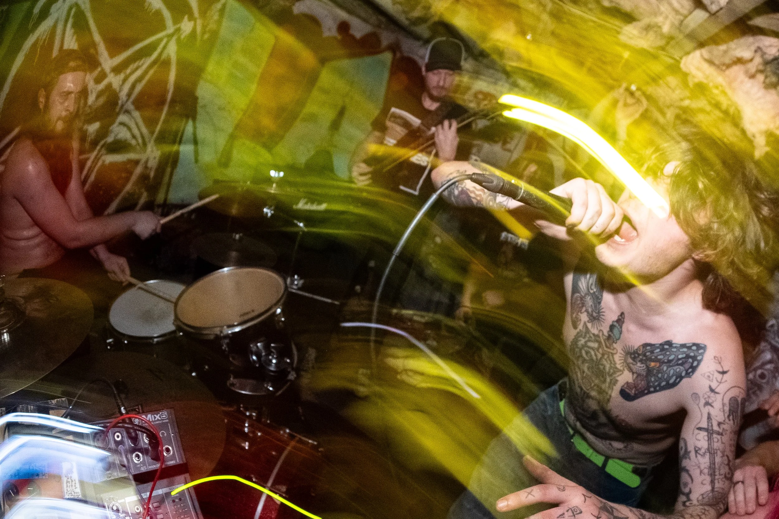 An energetic music performance in a dimly lit venue with yellow lighting effects. The shirtless vocalist with tattoos is singing into a microphone, and a drummer with long hair is playing in the background.