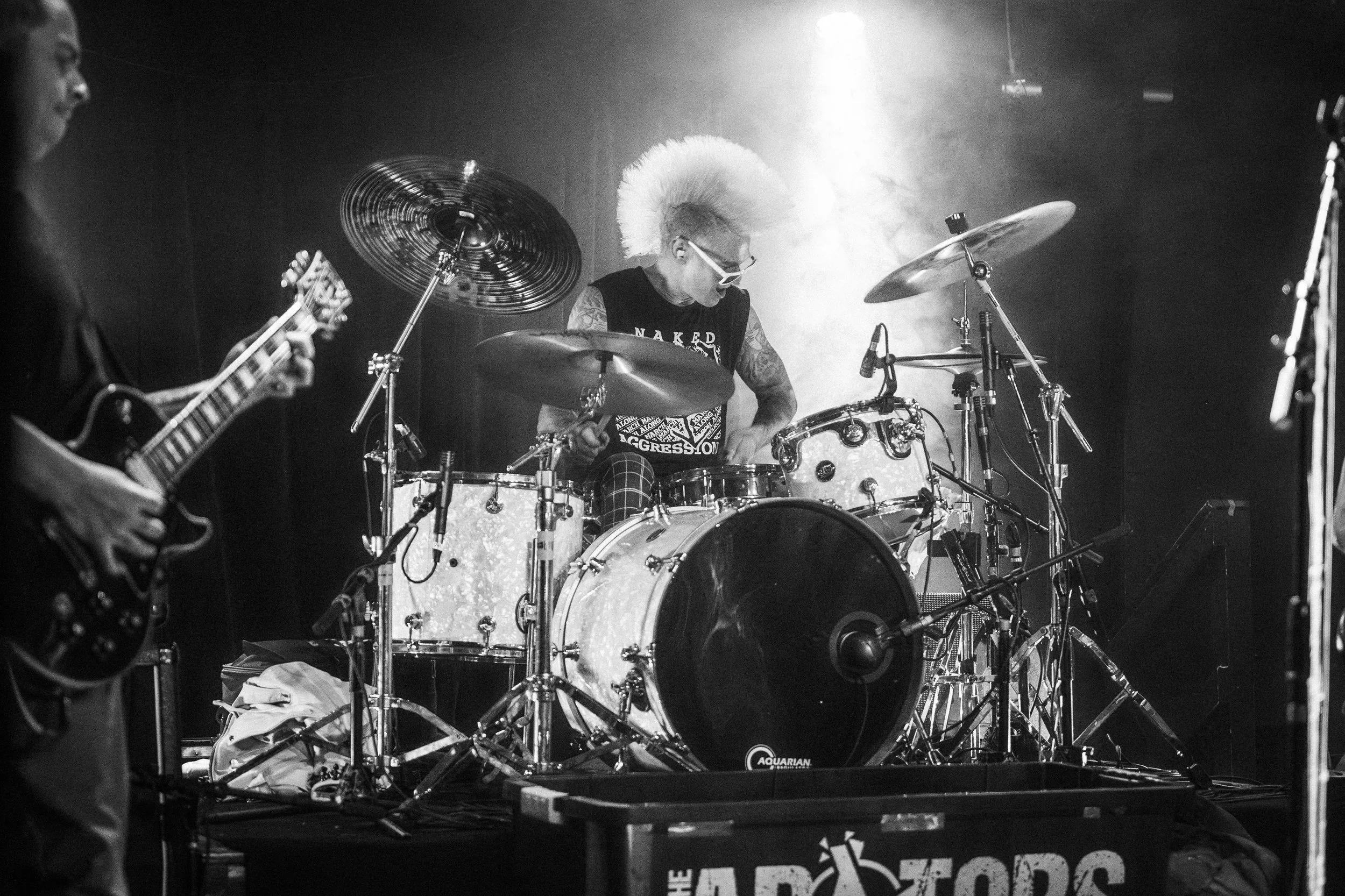 A musician with a mohawk playing drums on stage, with another musician playing guitar partially visible on the left, in a black and white photo.