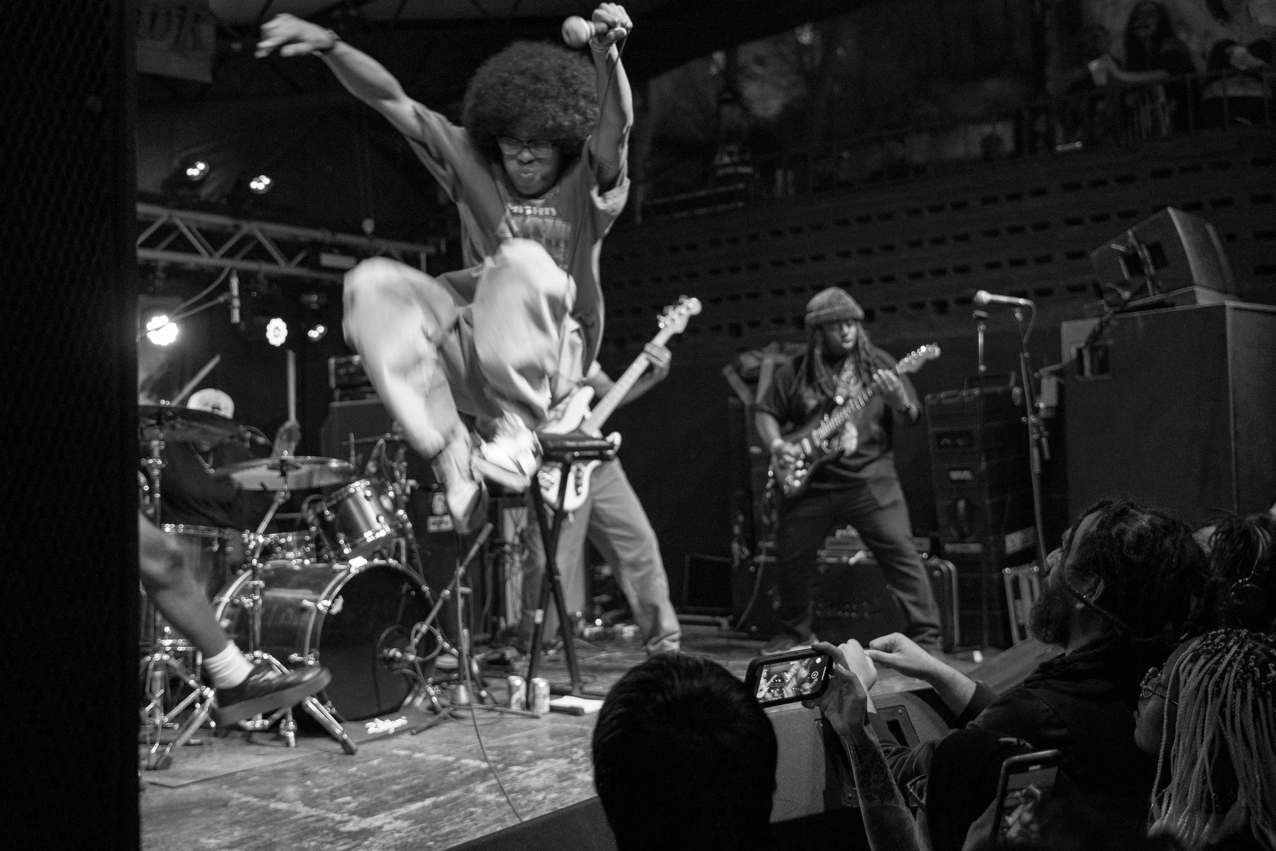 Two guitarists performing on stage at a concert, with one jumping in mid-air and the other playing guitar, in front of an audience.