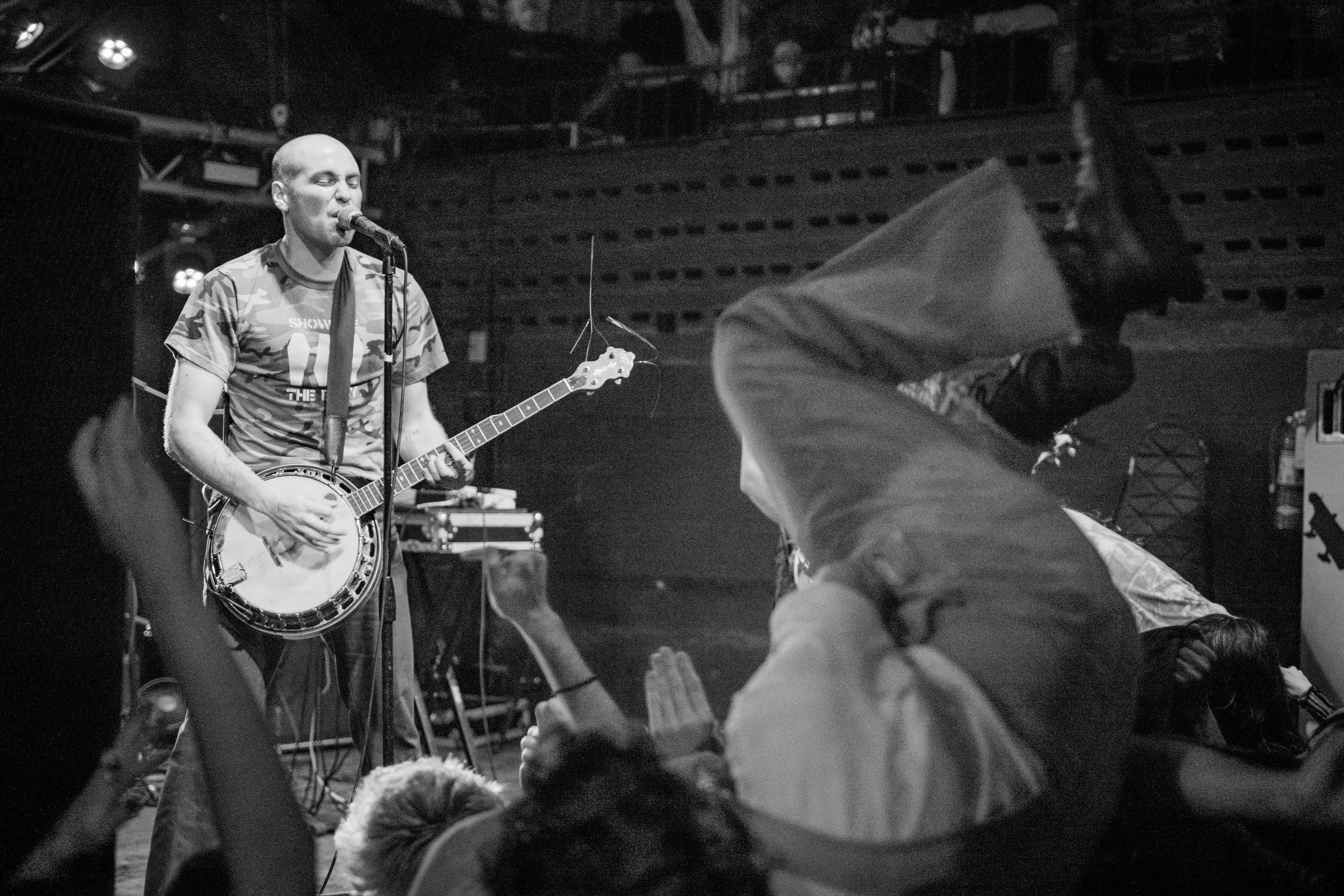 A musician performing on stage with a guitar and microphone, with an audience in front of him. The scene is in black and white, with some audience members raising their hands or wearing hooded clothing.