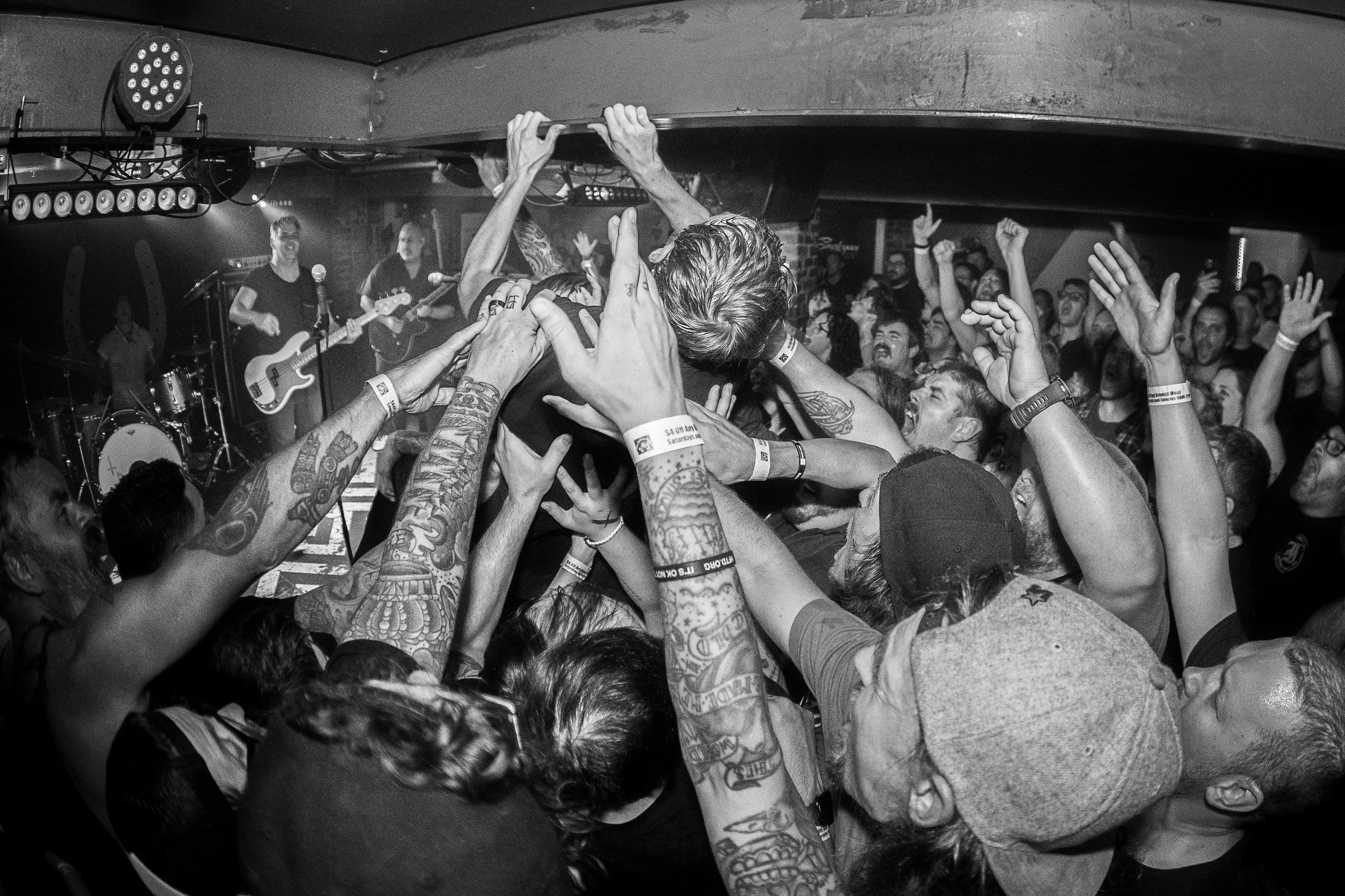 Crowd of people at a concert, crowd surfing and raising hands, band playing on stage in background, black and white photo.
