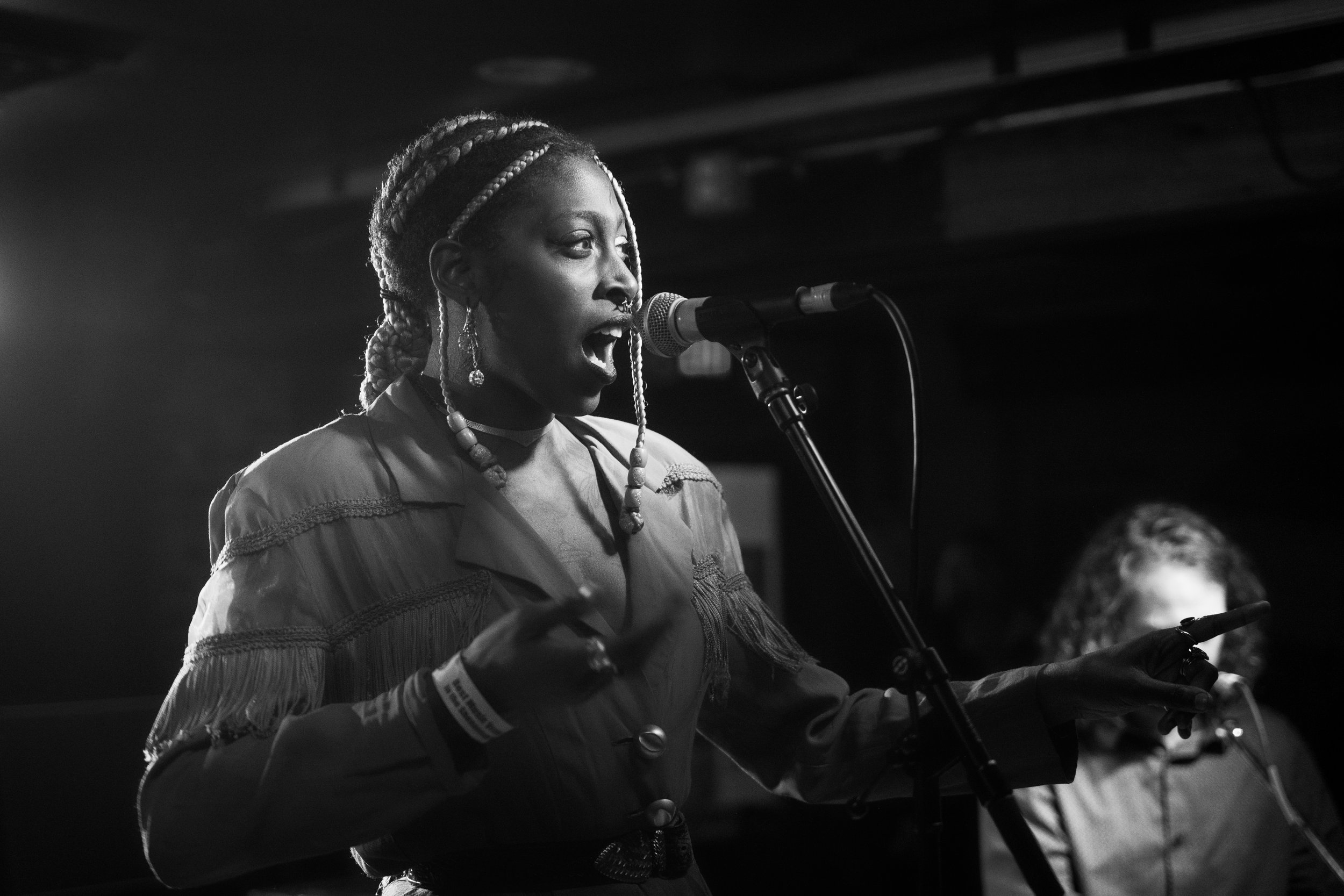 Black and white photo of a woman singing into a microphone with a person in the background.