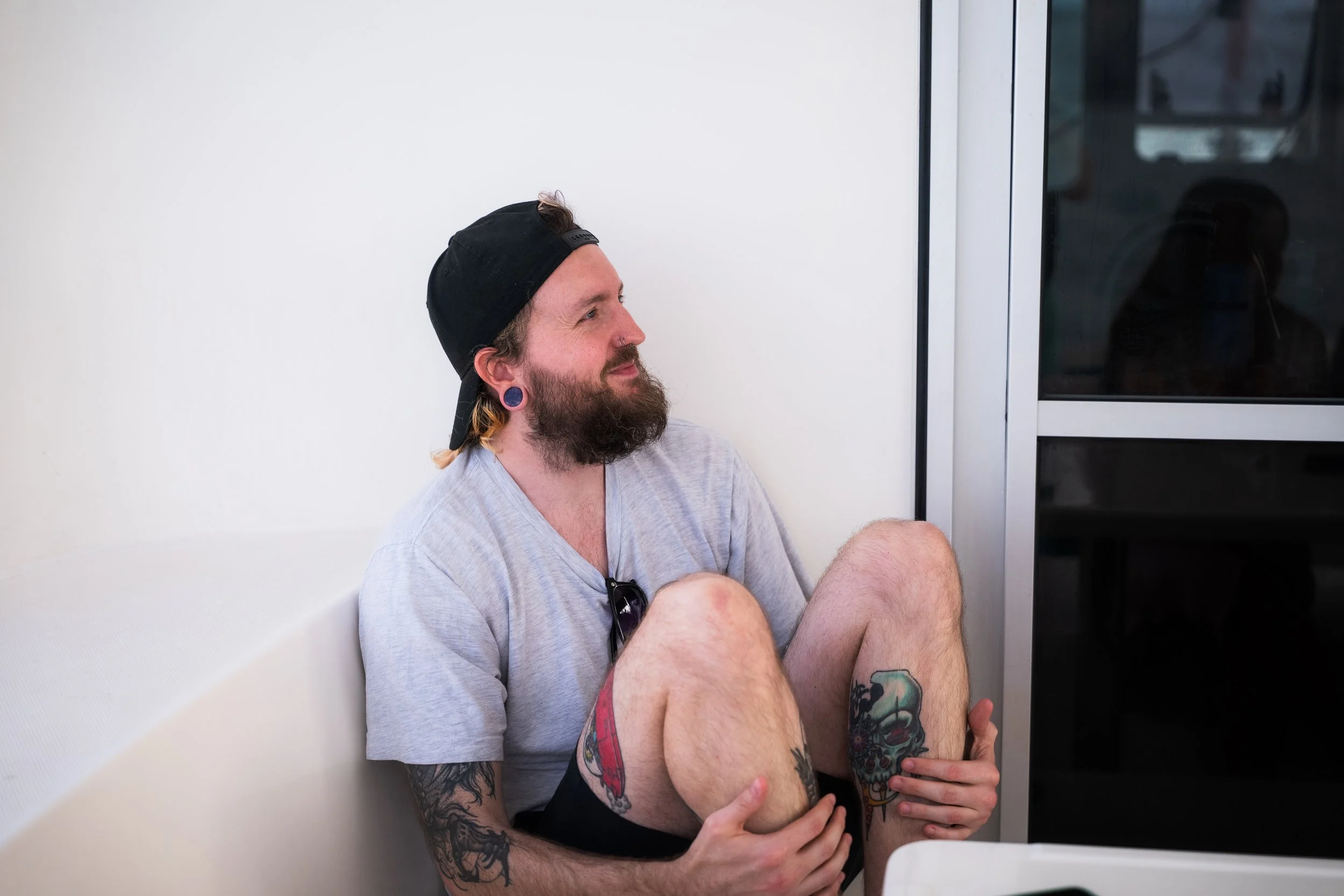 A man with a beard, tattoos, and wearing a black cap backwards, sitting on the floor near a white wall, holding his knees, with a partially visible window beside him.