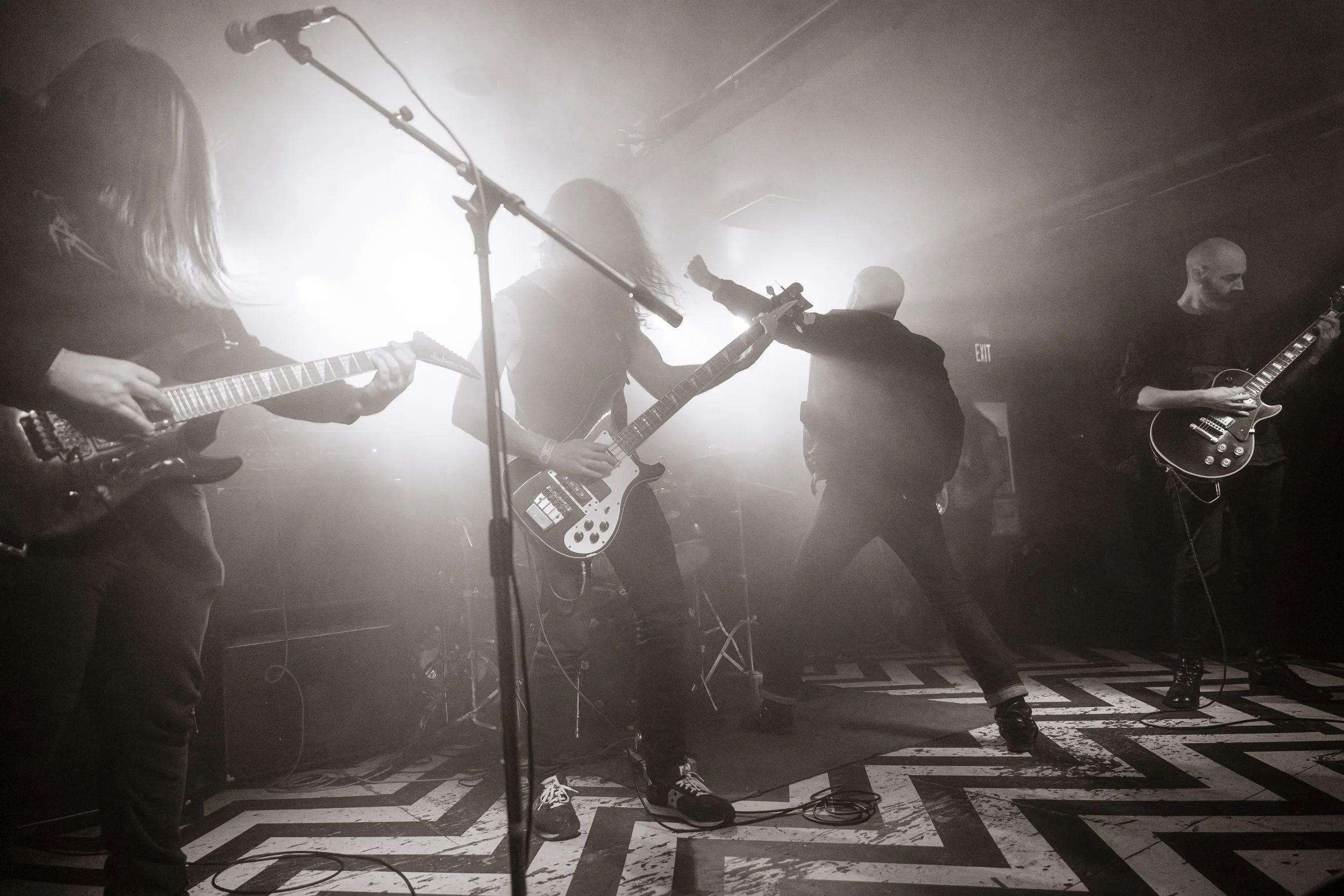 A band performing live on stage with four members playing electric guitars and a drummer. The scene is in black and white with bright backlighting creating a dramatic atmosphere.