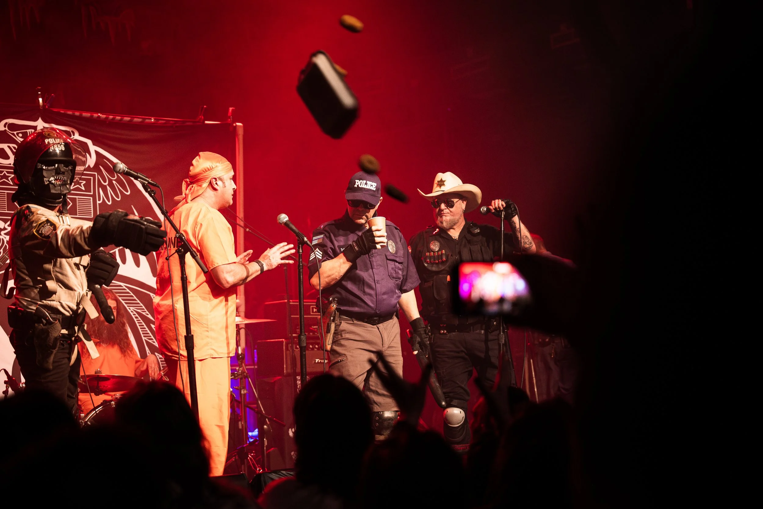 Musicians on stage performing at a concert, dressed as police officers and a prisoner, with one wearing a cowboy hat, amidst red stage lighting and an audience in front.