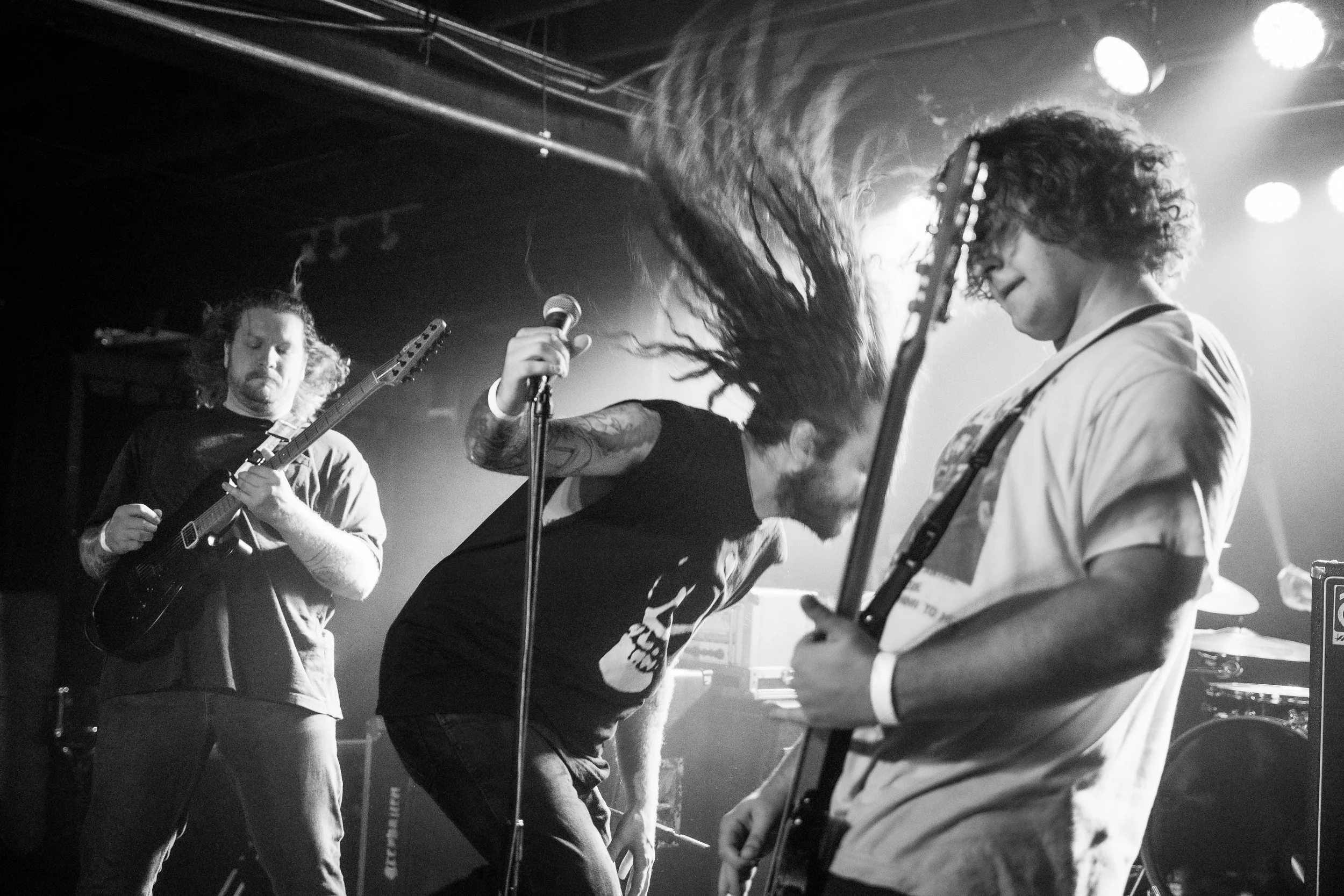 Black and white photo of three musicians performing on stage. The musician on the left is playing an electric guitar, the musician in the middle is singing into a microphone with long hair flying, and the musician on the right is playing another elec