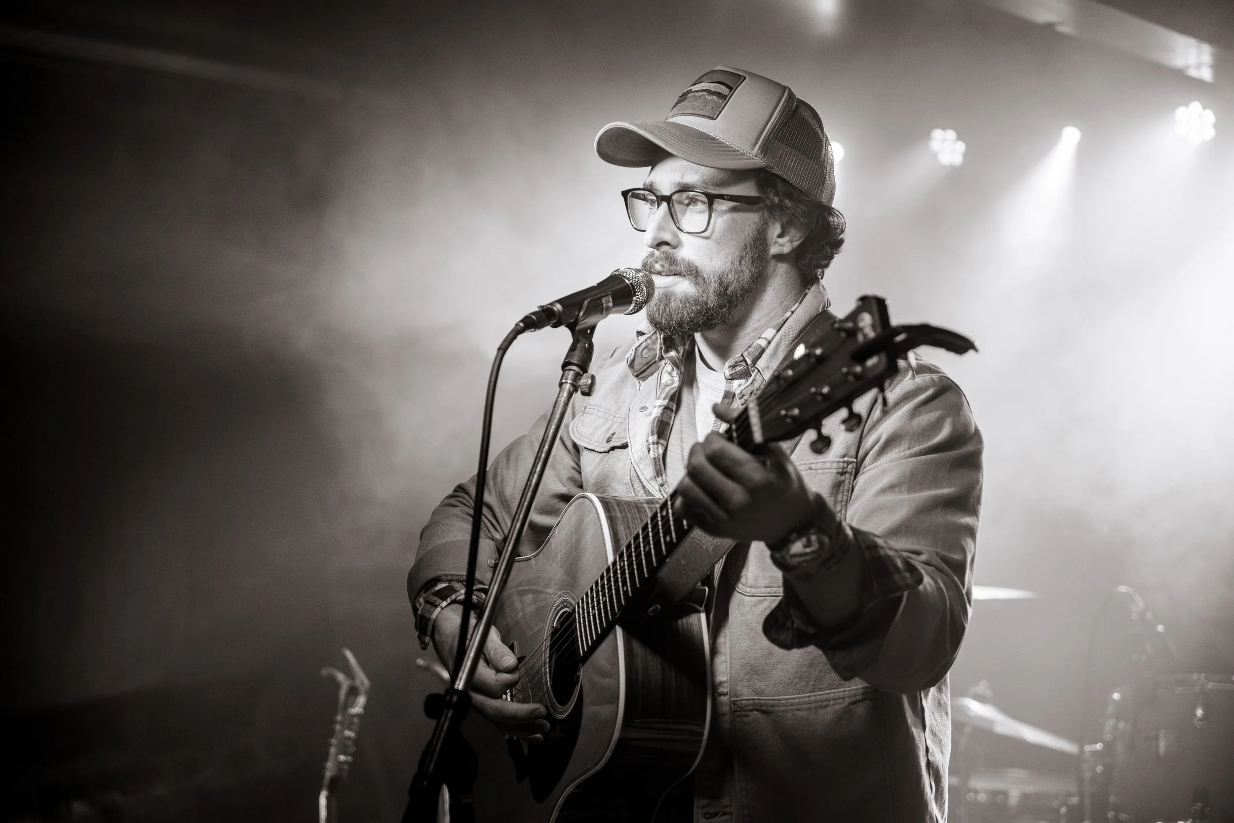 A man with glasses and a beard, wearing a cap and denim jacket, singing into a microphone while playing an acoustic guitar on stage with spotlights overhead