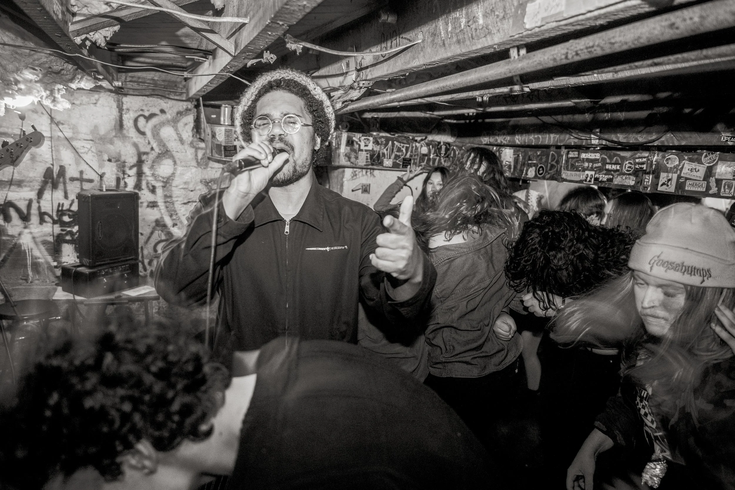 A man with curly hair, glasses, and a beard singing into a microphone, pointing towards the camera, in a crowded, graffiti-covered basement with people dancing around him.