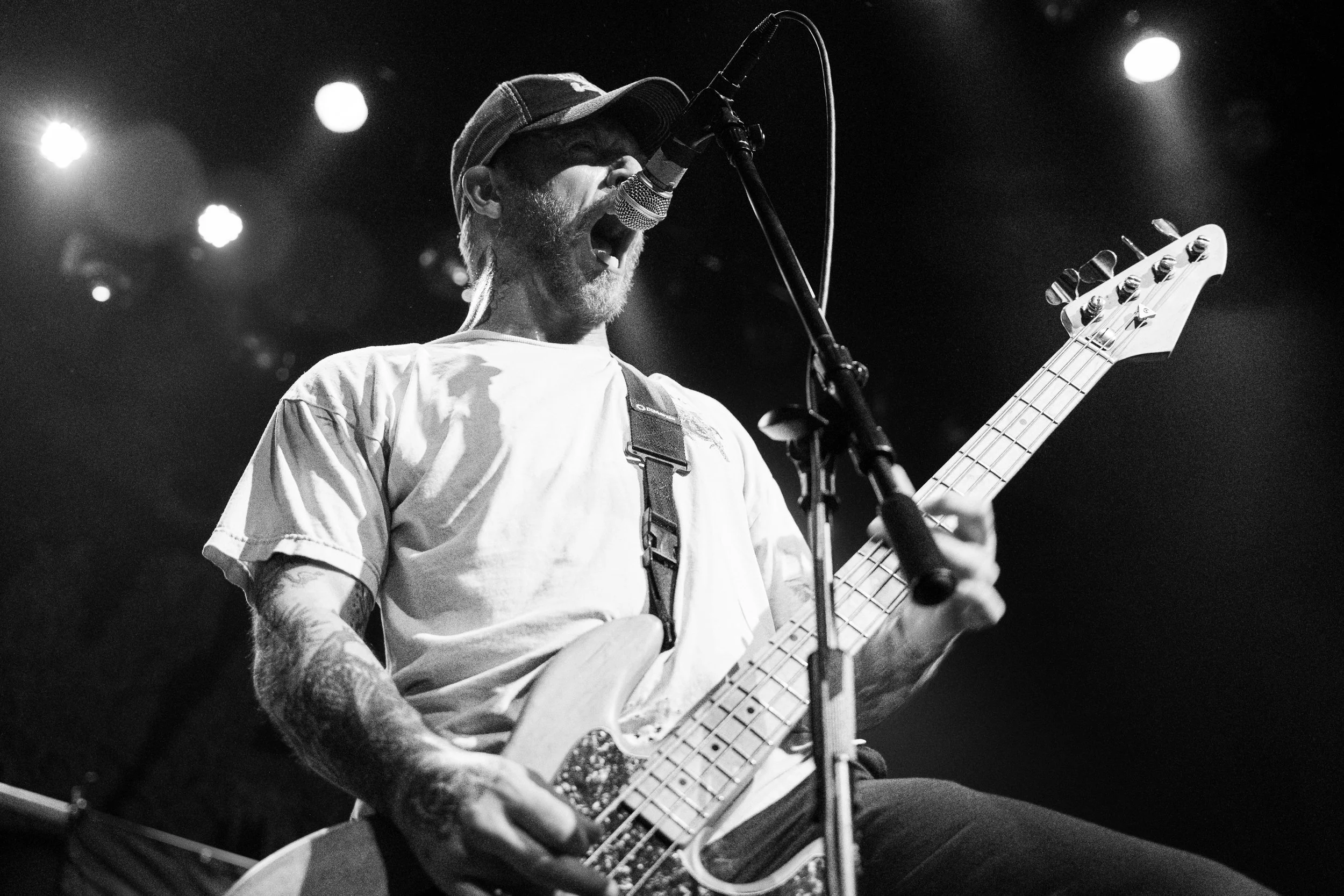 A male musician singing into a microphone while playing an electric bass guitar on stage, wearing a baseball cap and a light-colored t-shirt, with stage lights in the background.