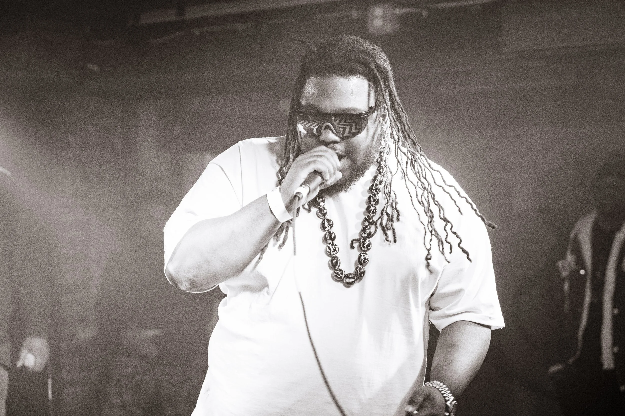 A man with dreadlocks wearing sunglasses, a white shirt, a large beaded necklace, and a bracelet, is holding a microphone and performing on stage with a dark background.