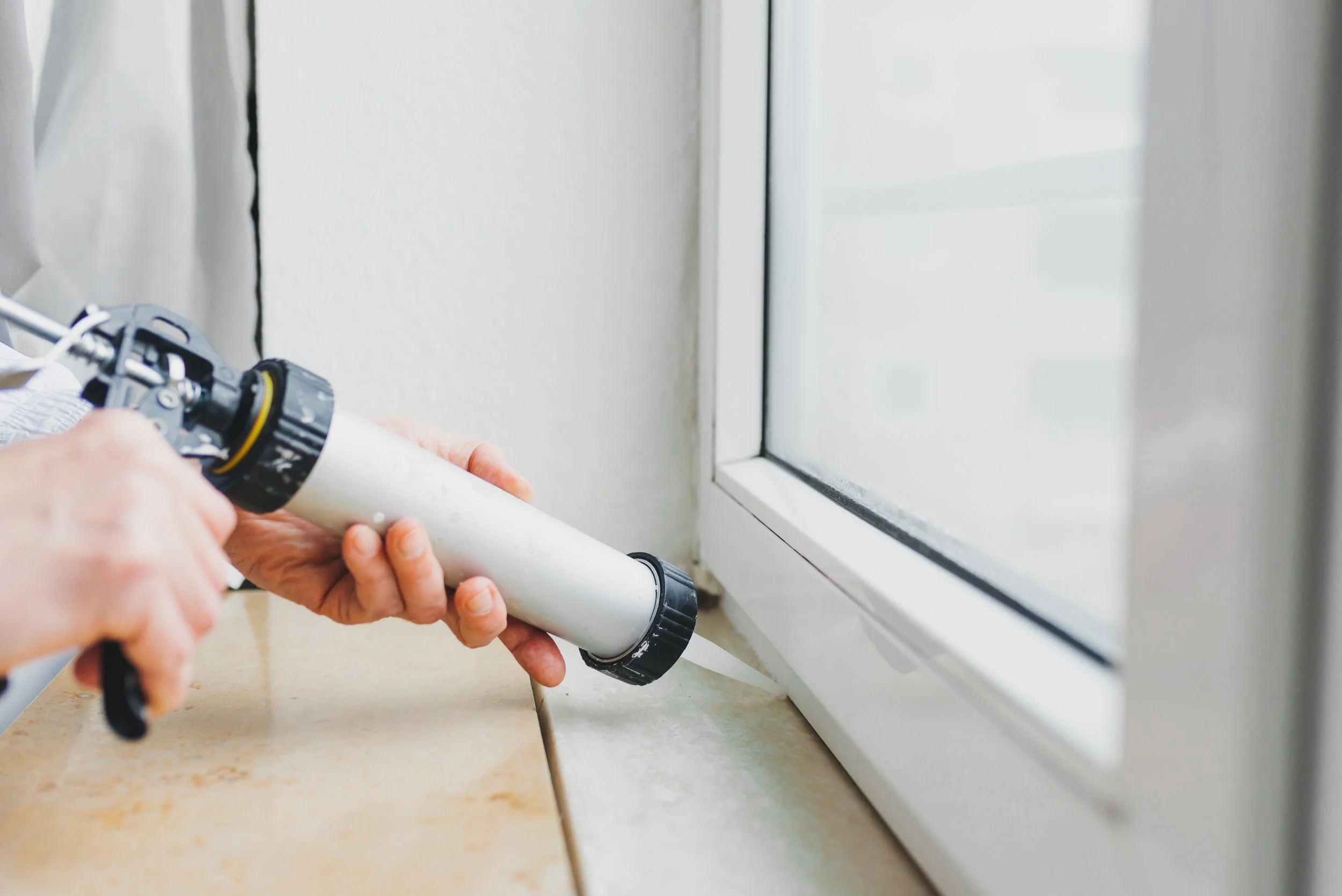 Person applying sealant around a window frame with a caulking gun.