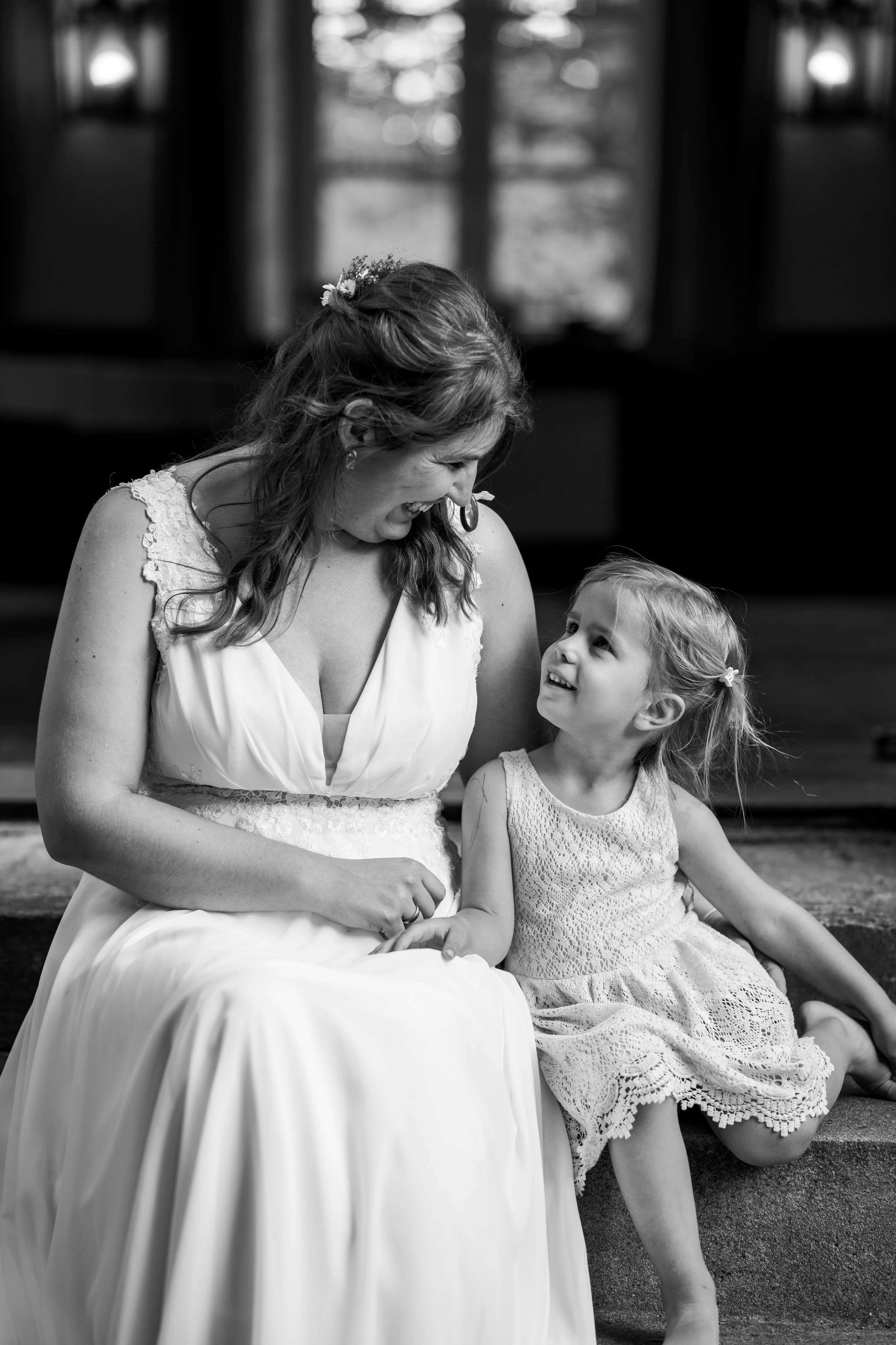 Schwarz-weiß-Foto von einer lachenden jungen Frau in einem Brautkleid, die mit einem kleinen Mädchen sitzt, das ein Sommerkleid trägt, im Hintergrund Fenster mit Vorhängen.