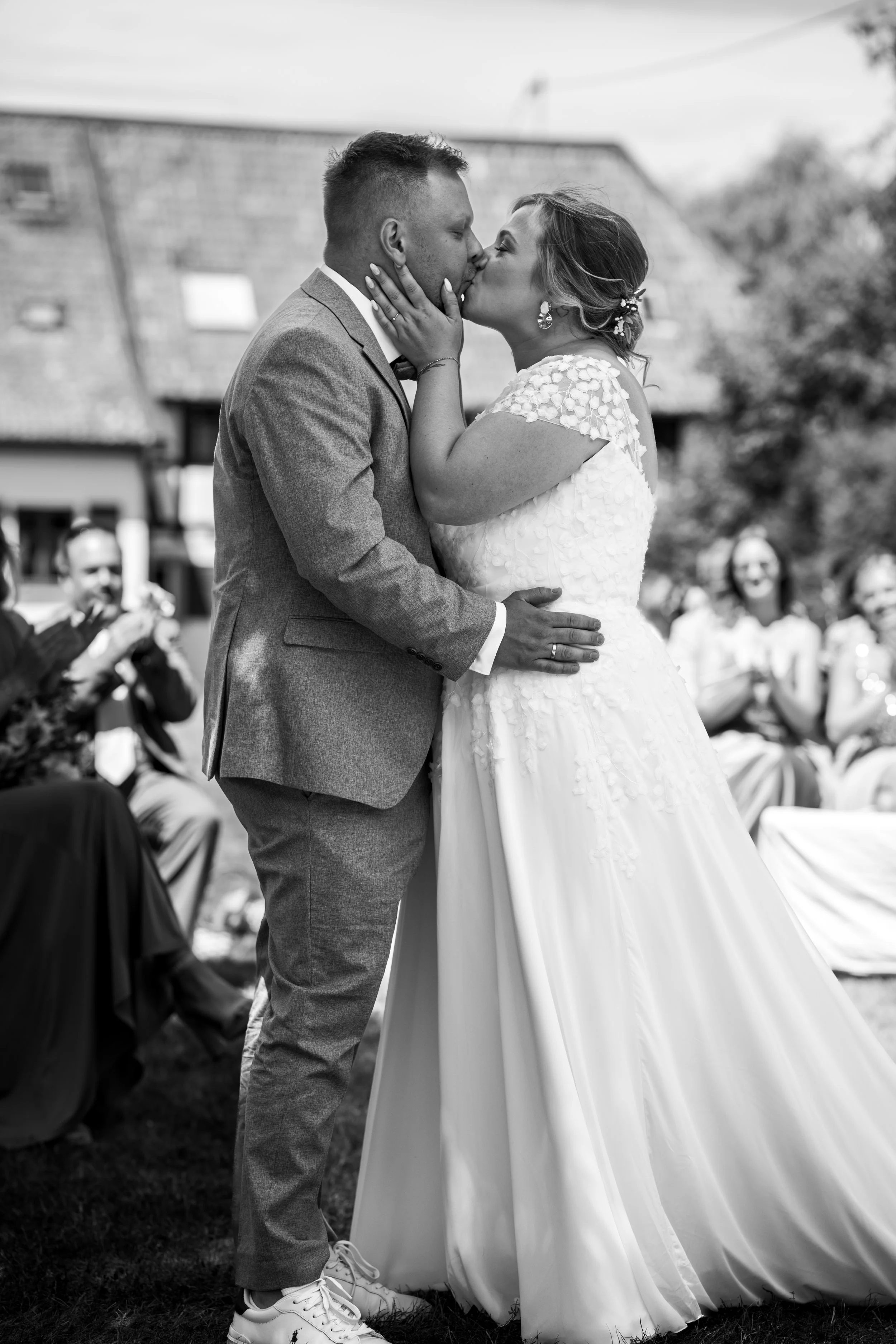 Braut und Bräutigam küssen sich bei ihrer Hochzeit im Freien, während Gäste ihnen zusehen und applaudieren.