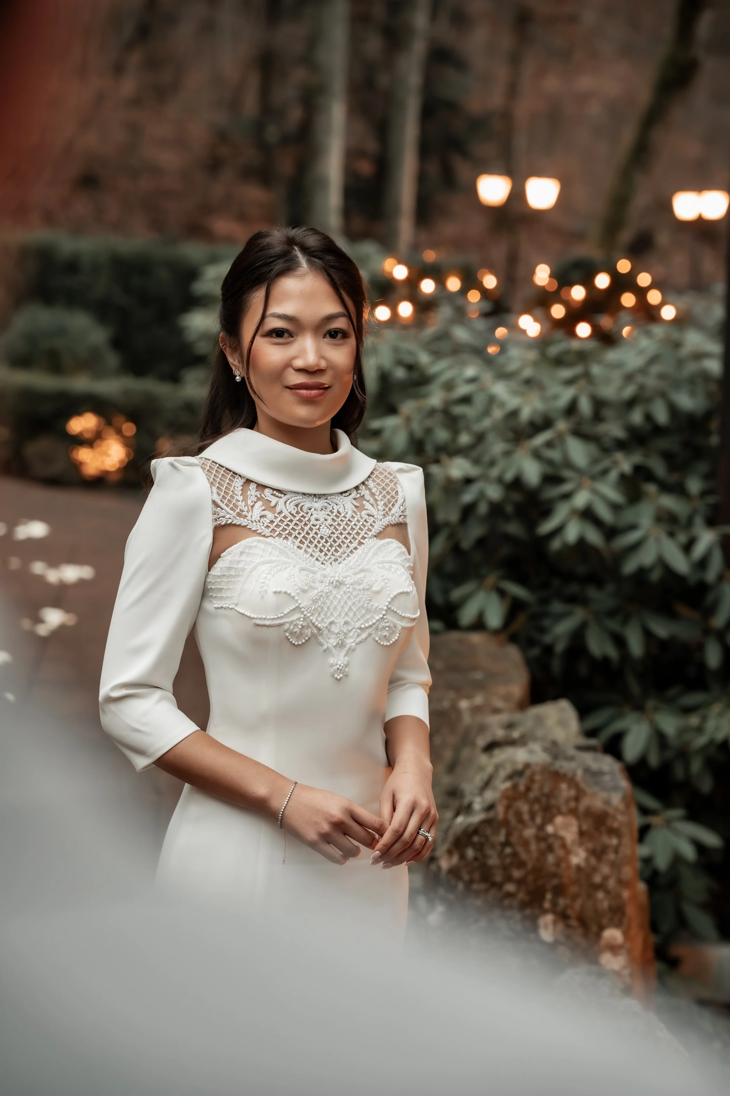 Eine junge Frau in einem weißen Kleid mit Perlen- und Spitzendetails steht in einem Garten mit grünen Büschen und Hintergrundlichtern.