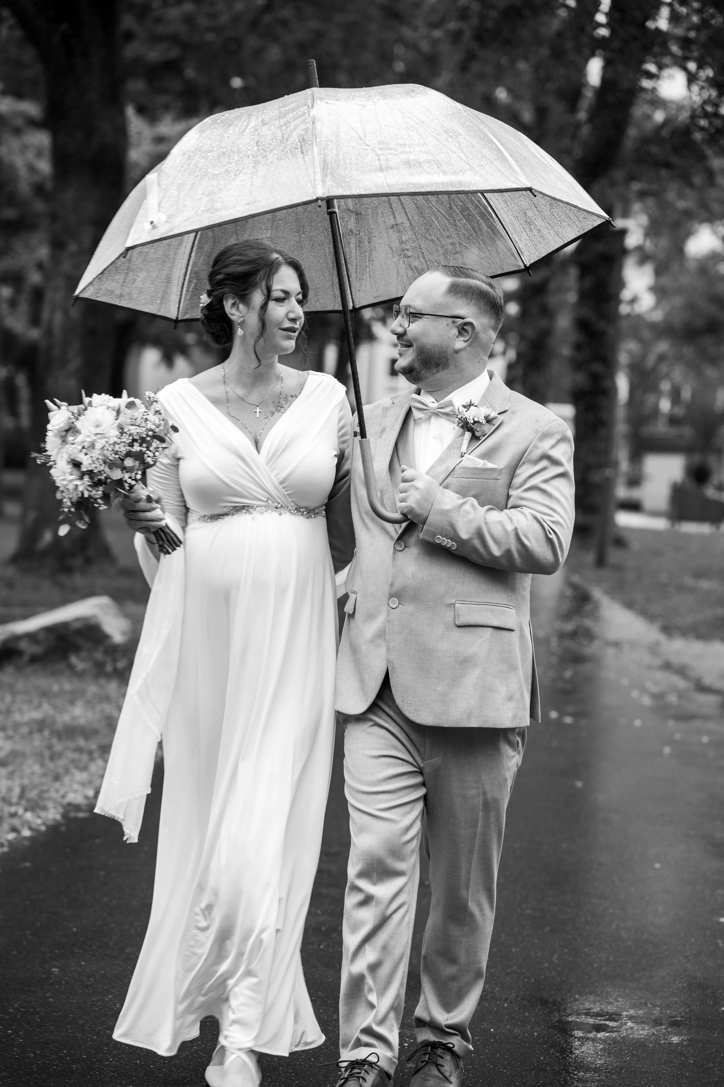 Ein brautpaar bei Hochzeit im Regen, der Bräutigam hält einen Regenschirm über die Braut, beide lächeln sich an, im Hintergrund Bäume.