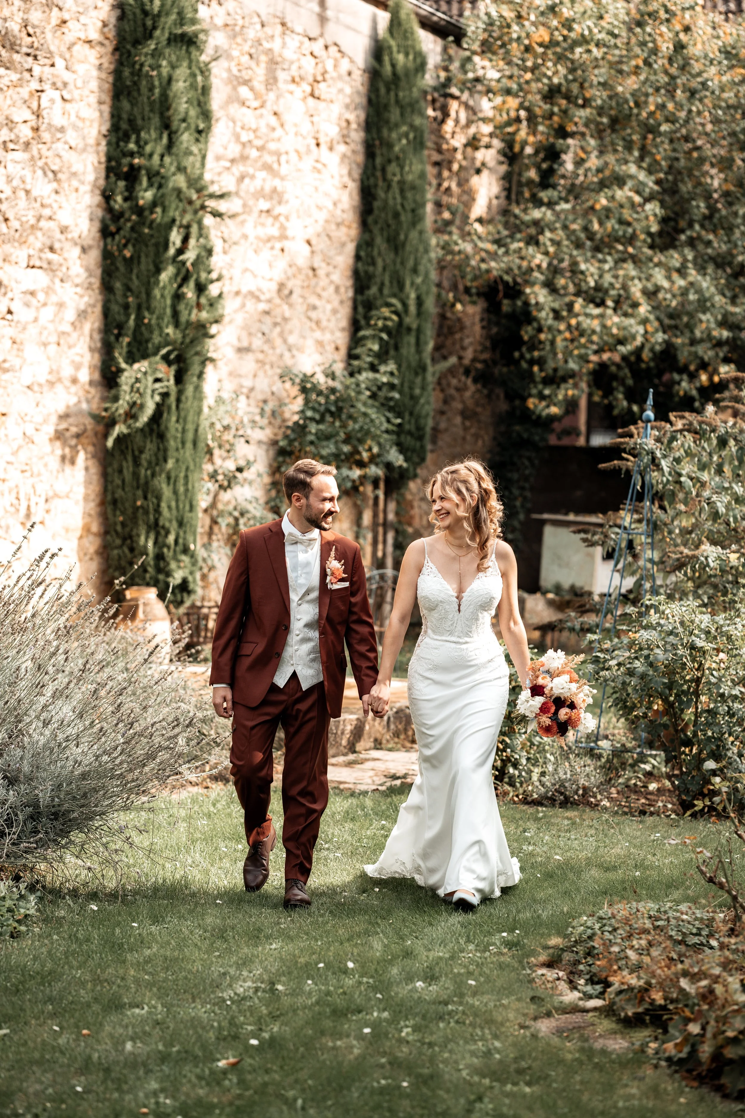 Ein Brautpaar läuft Hand in Hand durch einen Garten, lächelt sich an und trägt Hochzeitskleidung.