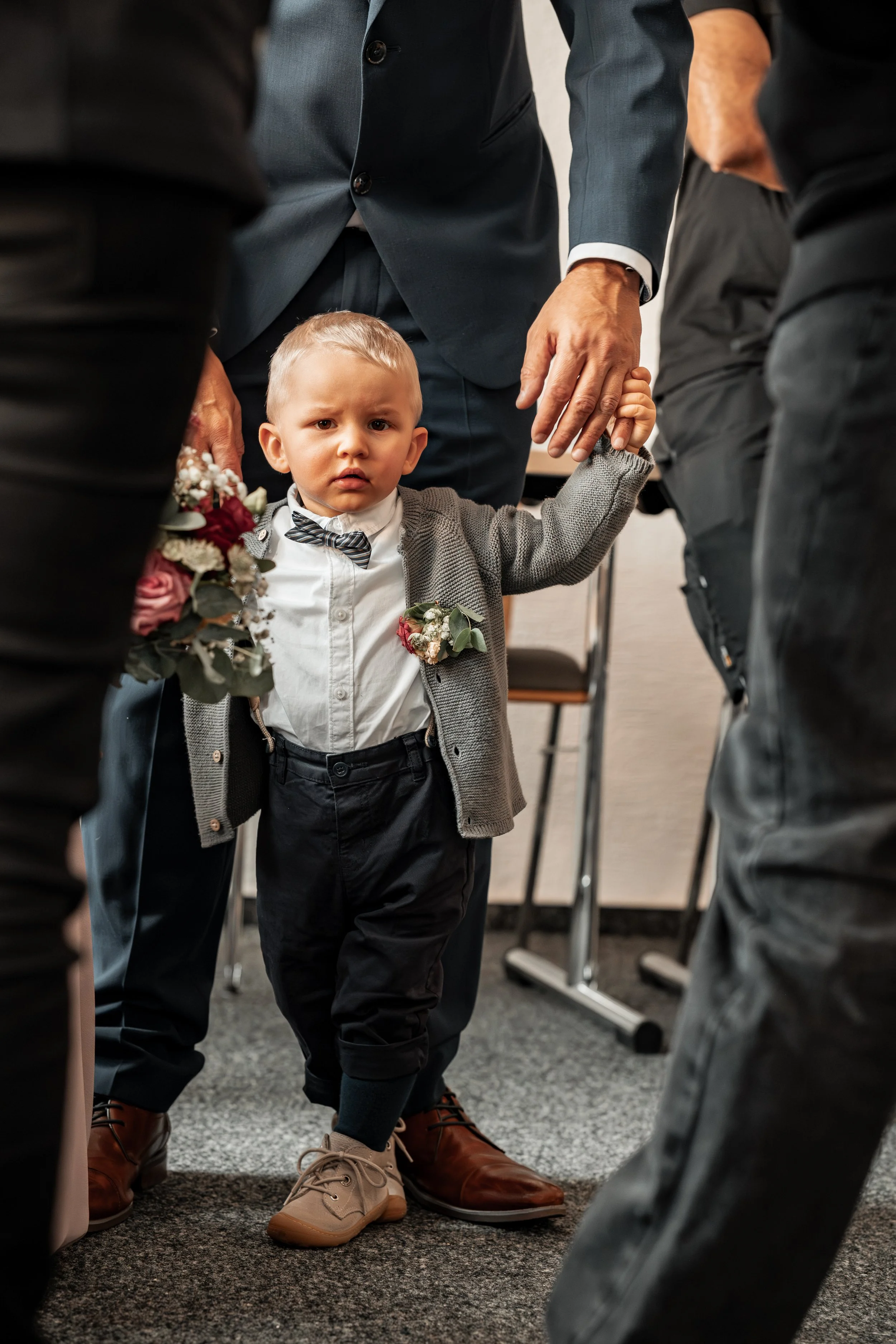 Ein kleiner Junge in formeller Kleidung, der von Erwachsenen auf einem Boden in einem Raum gehalten wird, wahrscheinlich bei einer Zeremonie oder einem besonderen Anlass.