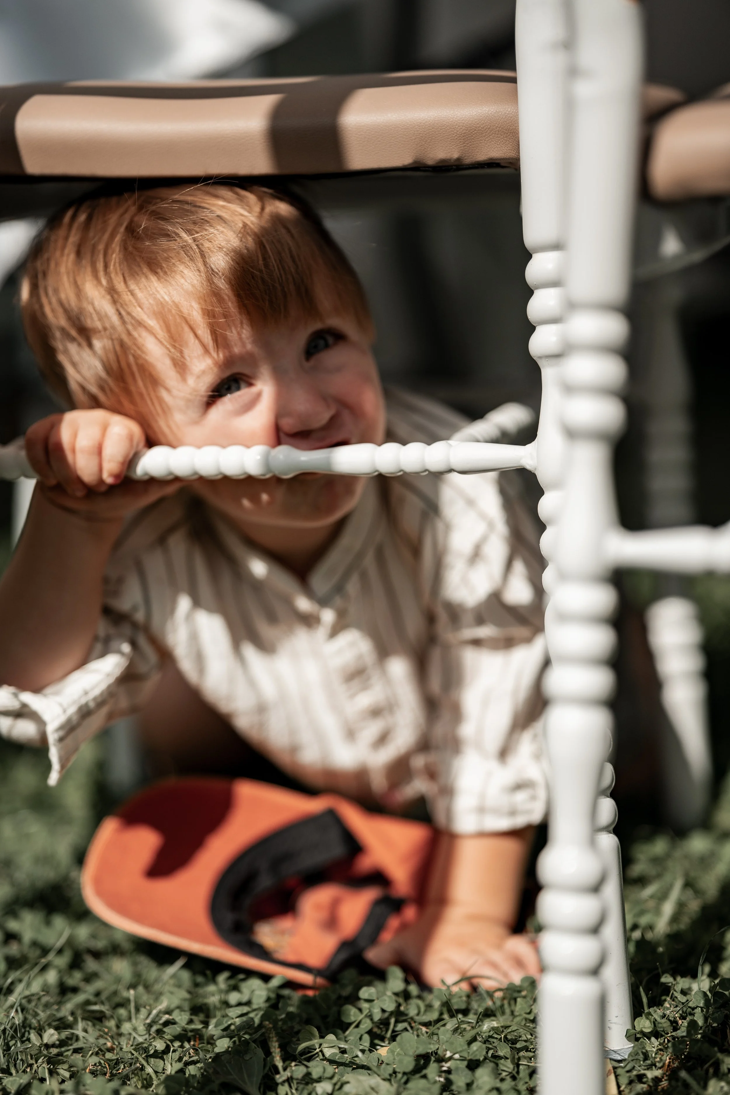 Ein kleiner Junge kriecht unter einem weißen Holzstuhl auf grünem Gras, trägt ein gestreiftes Hemd und orange Gurt, und lächelt.