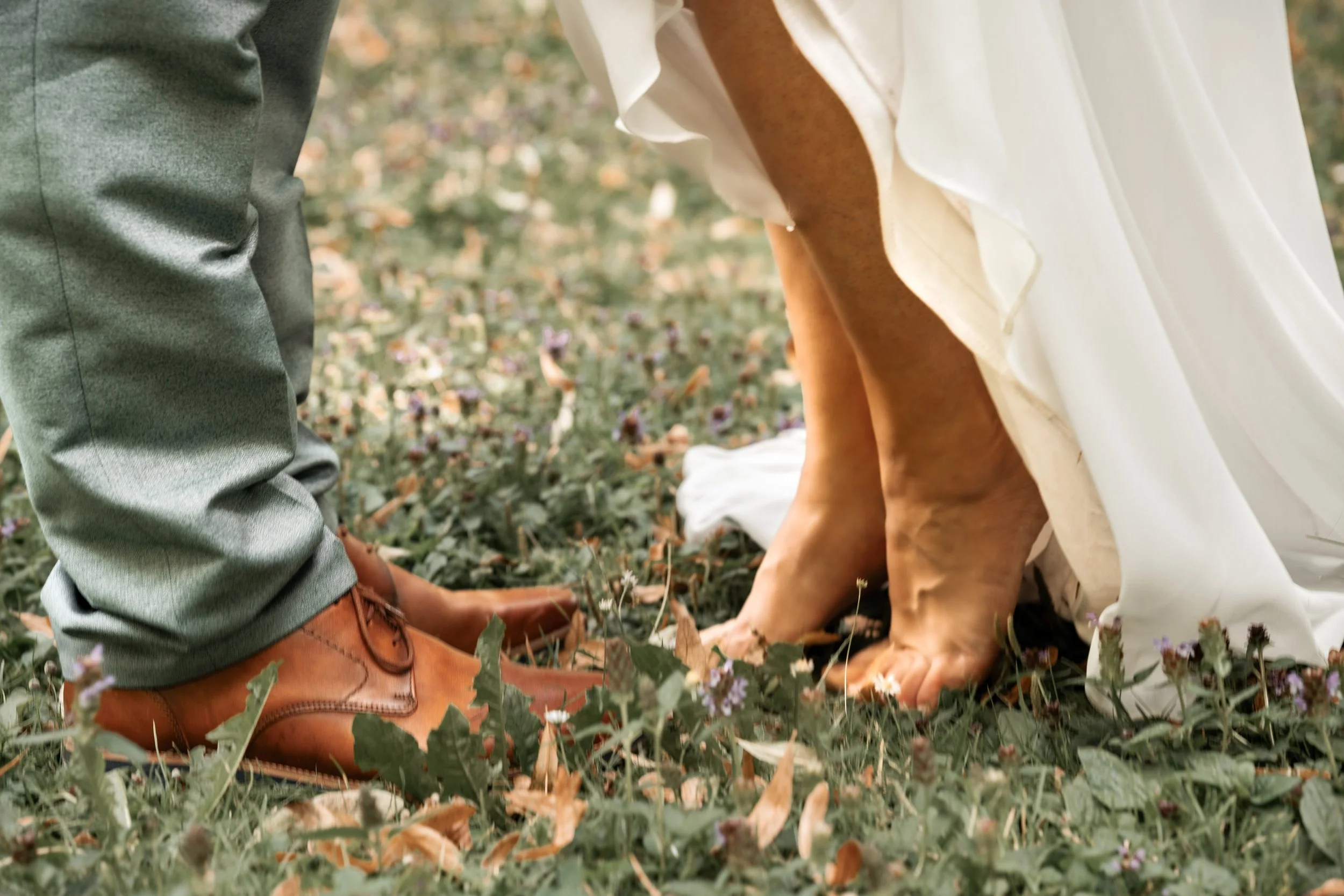 Ein Brautpaar bei der Hochzeit, die Füße eines Paares stehen auf dem Rasen, mit Blüten und Blättern umgeben, die Braut trägt ein weißes Kleid und barfuß, der Bräutigam trägt braune Schuhe und einen grauen Anzug.