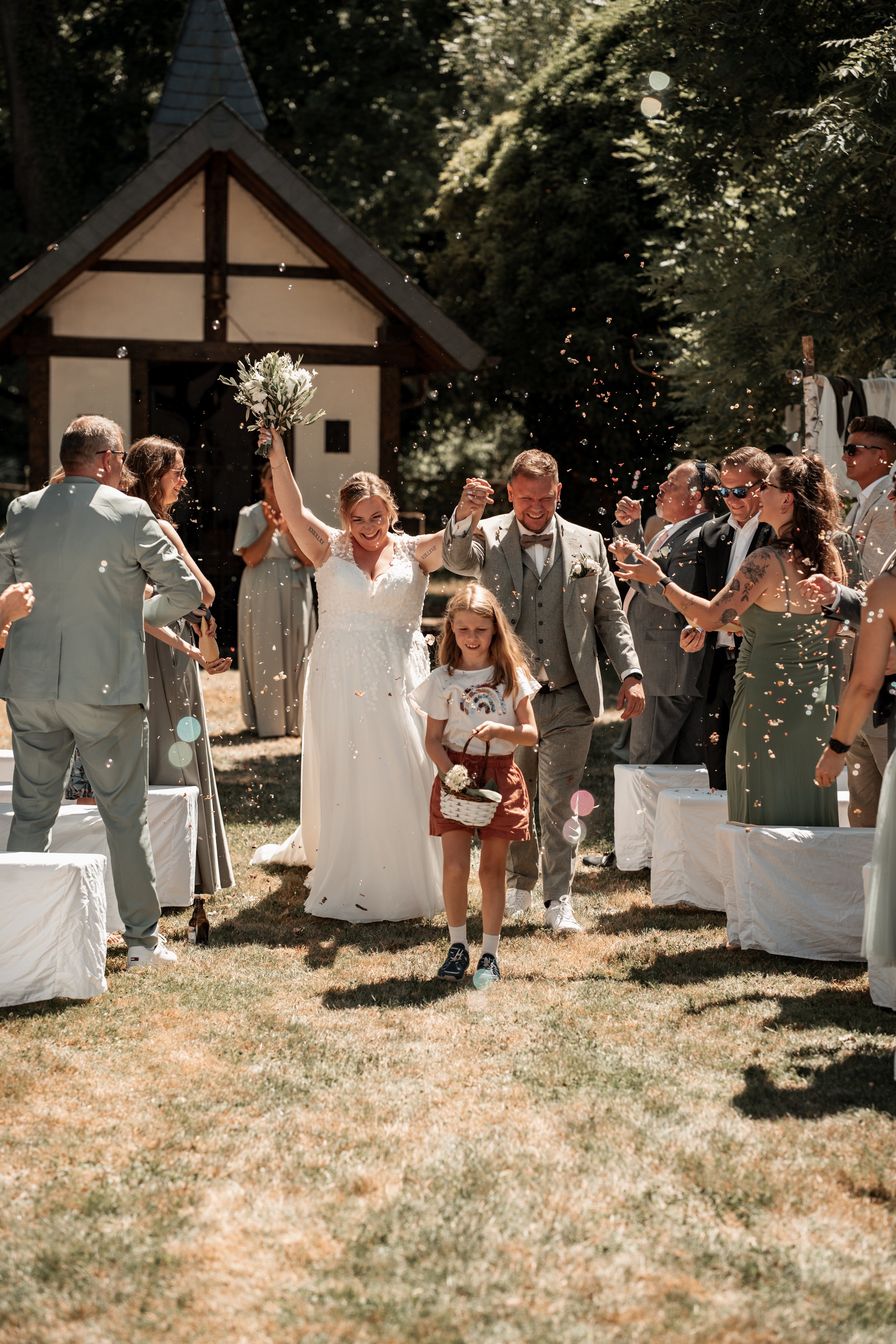 Ein Frischvermähltes Paar läuft lachend durch eine Menschenmenge bei einer Hochzeitsfeier im Freien, umgeben von Freunden und Familie, mit Papierkonfetti in der Luft.