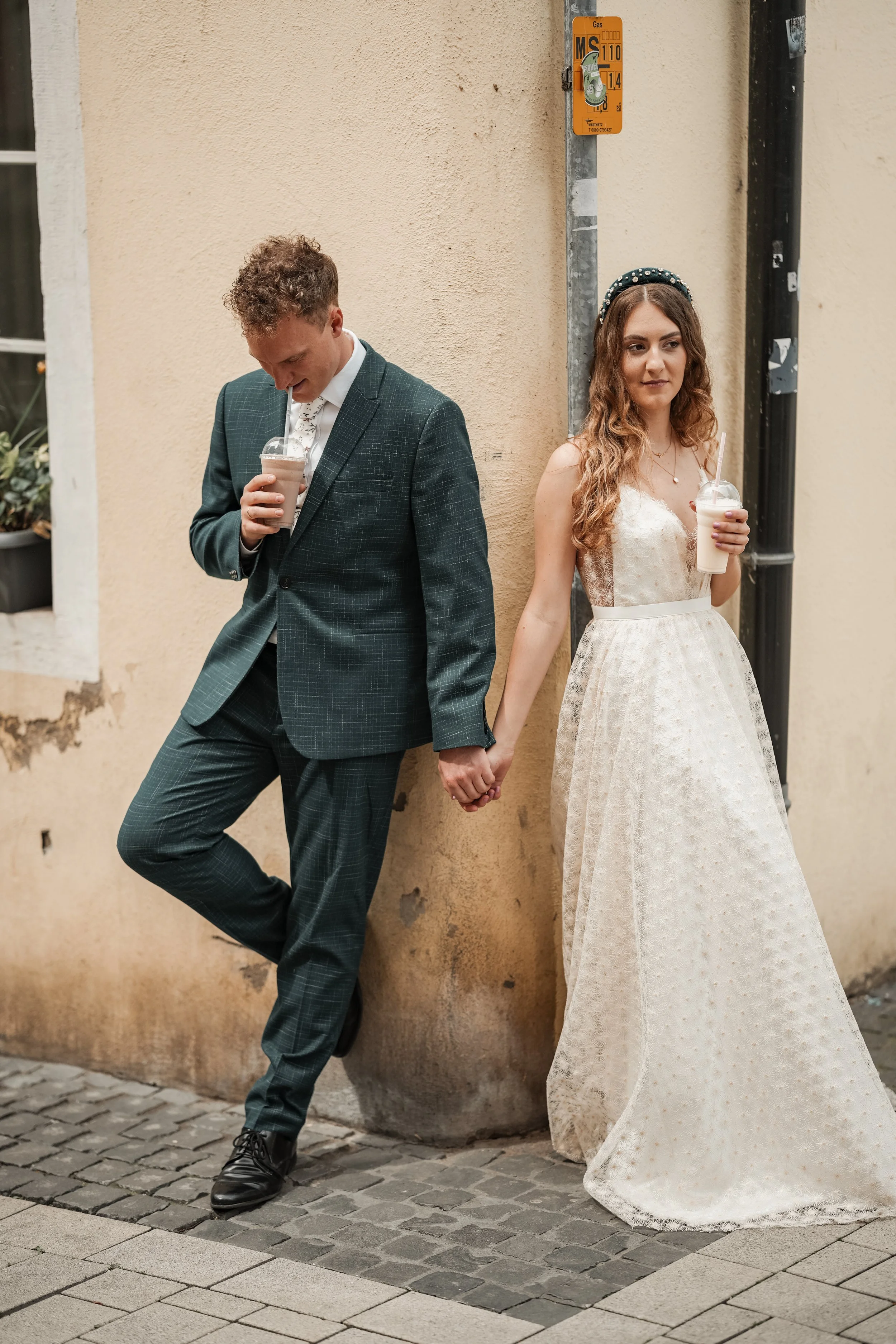 Ein Paar, das einander an einem Straßenrand die Hand hält, während sie Getränke trinken. Das Mädchen trägt ein weißes Hochzeitskleid und ein Haarband, der Junge trägt einen dunkelgrauen Anzug. Es sieht aus wie eine Hochzeit oder ein besonderer Anlass