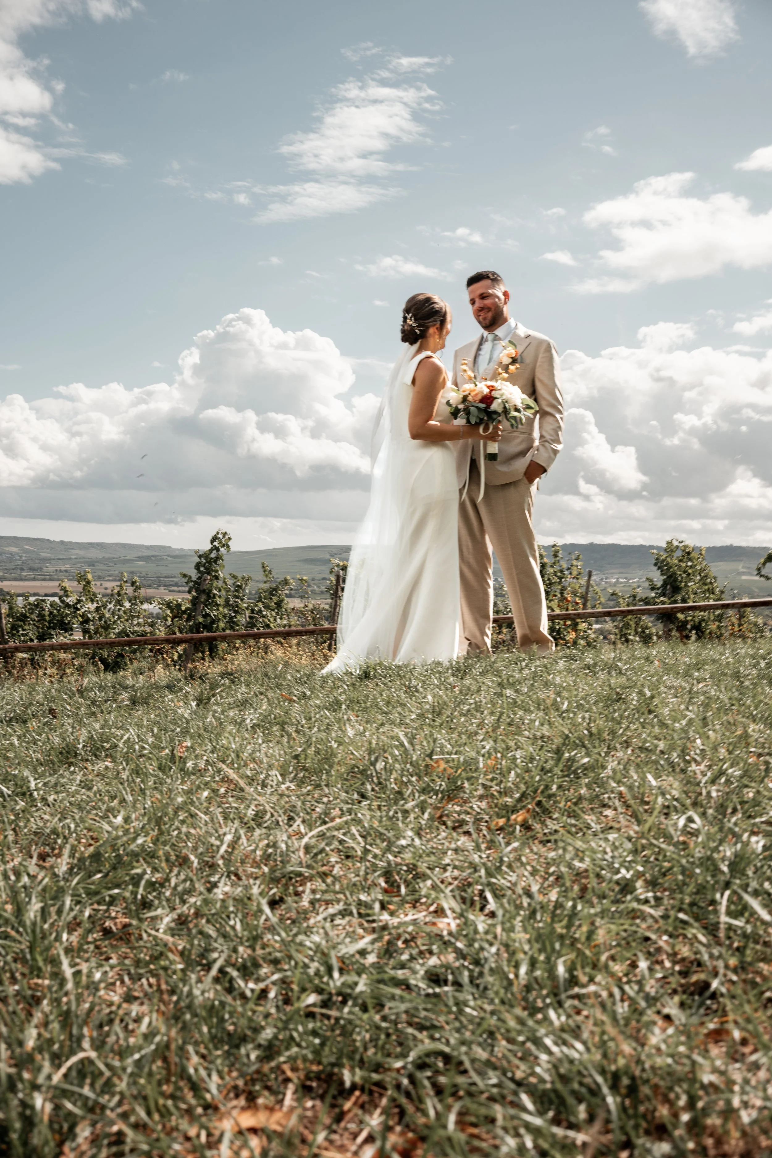 Braut und Bräutigam bei Hochzeit im Freien, stehende auf grünem Rasen, hinter ihnen Berge und Wolken im Himmel, Braut hält Blumenstrauß, Freundlichkeit zwischen den beiden erkennbar.
