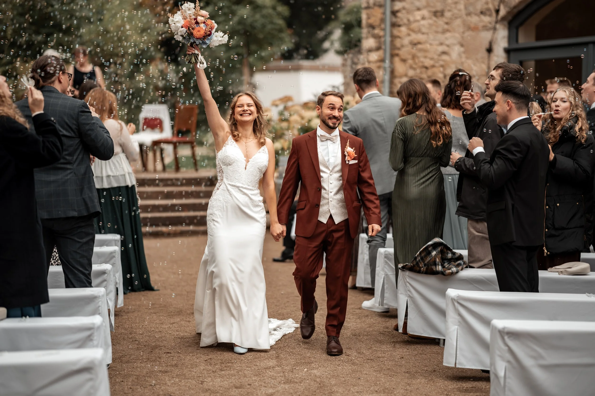 Ein glückliches Brautpaar läuft bei ihrer Hochzeit Hand in Hand durch eine Gruppe von Freunden und Familie, während sie ihre Hochzeit feiern, mit Konfetti und Blumen.