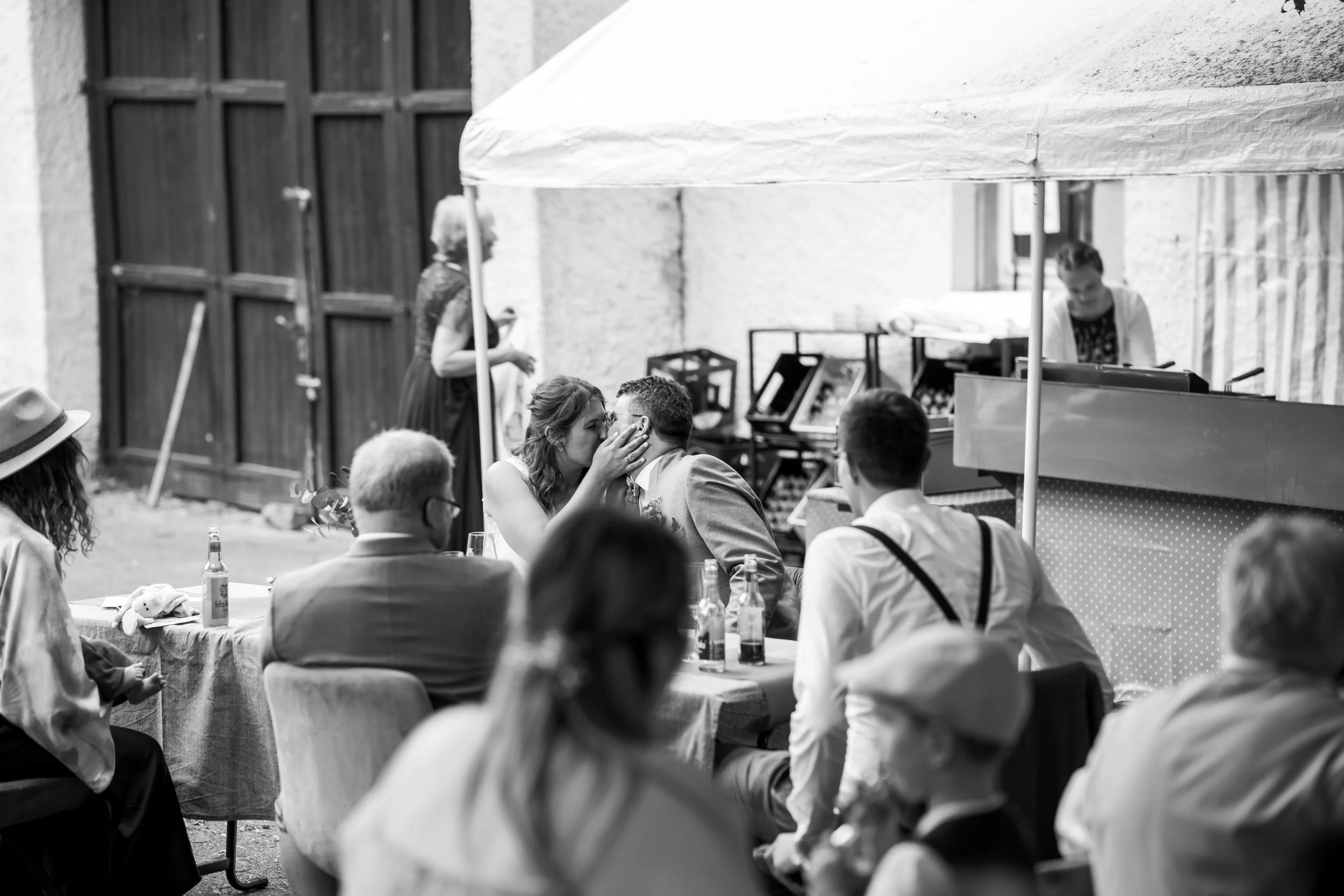 Ein schwarzes und weißes Foto von einer Hochzeitsfeier im Freien. Ein Brautpaar küsst sich, während die Gäste um sie herum sitzen, Getränke trinken und sich unterhalten. Ein Stand mit einer Verkäuferin ist im Hintergrund sichtbar.
