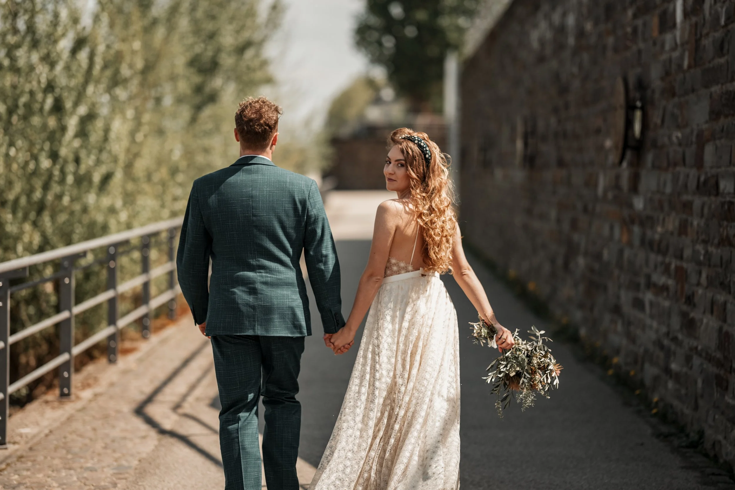 Ein Brautpaar hält sich an den Händen, während es an einer Steinmauer entlanggeht. Die Frau trägt ein weißes Hochzeitskleid und hält einen Blumenstrauß, der Mann trägt einen dunklen Anzug. Es ist sonnig und die Umgebung ist grün mit Bäumen.