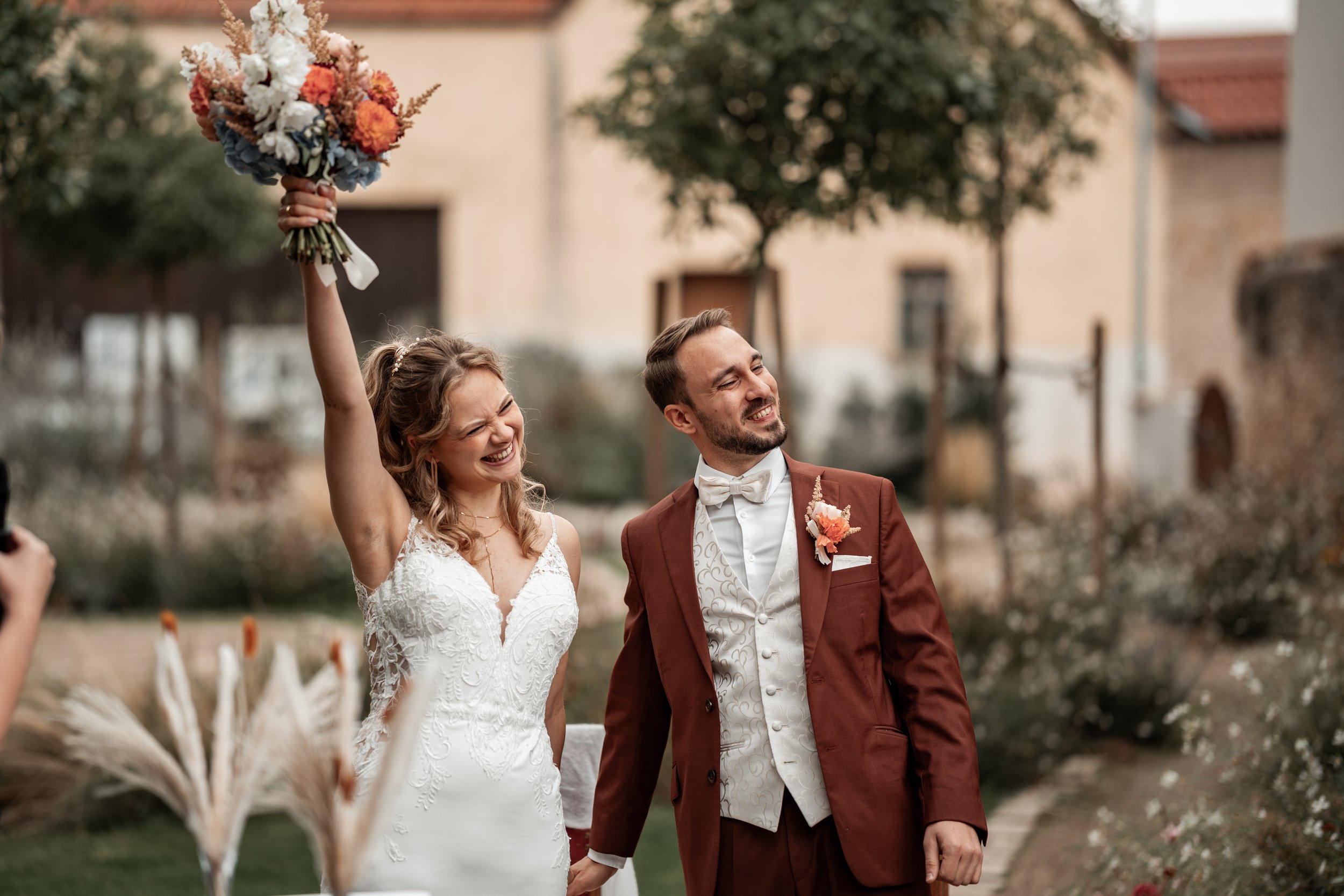 Ein glückliches Brautpaar bei ihrer Hochzeit draußen, sie hält den Brautstrauß hoch, er trägt einen braunen Anzug, sie trägt ein weißes, elegantes Brautkleid.