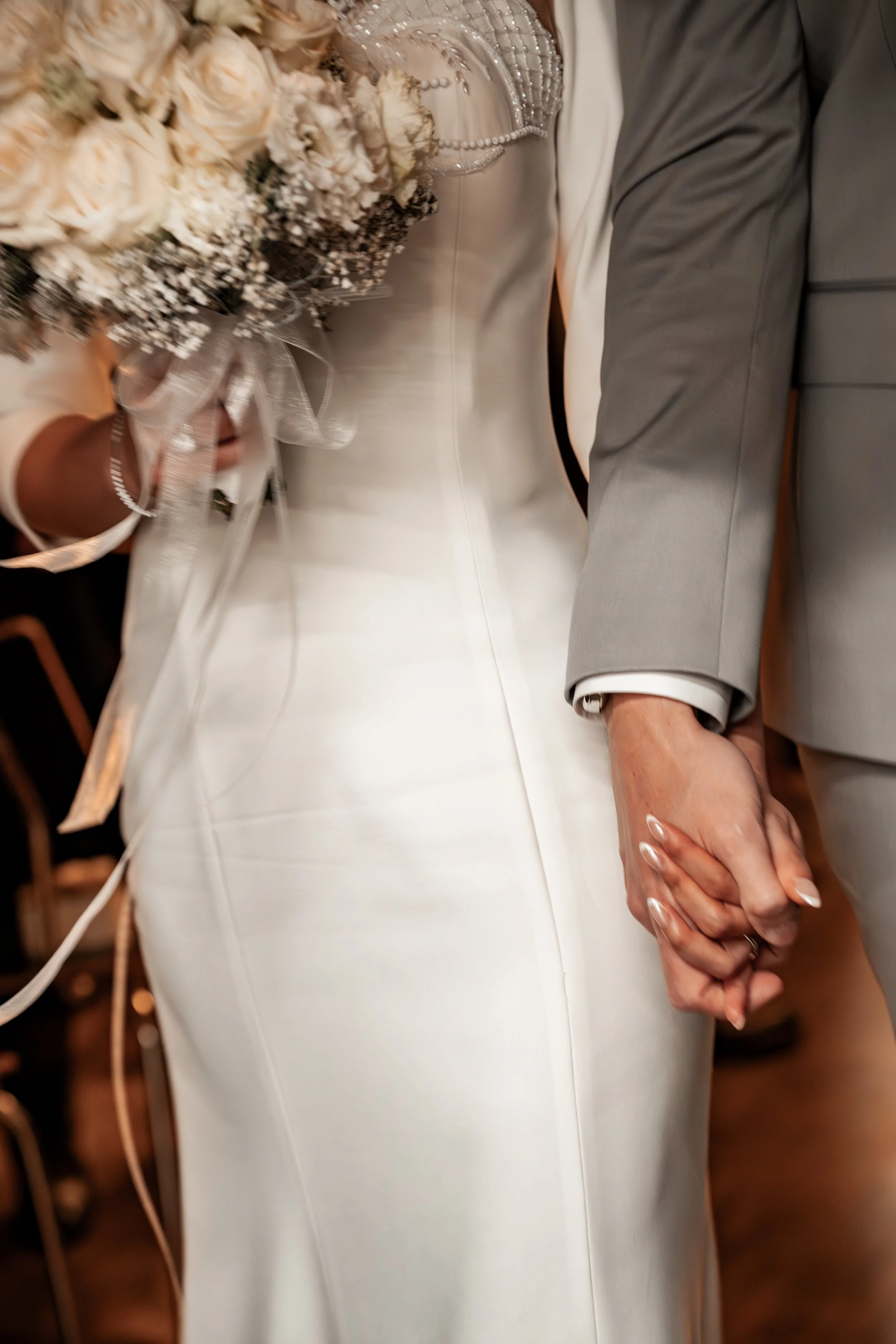 Ein Brautpaar bei der Hochzeit hält sich an den Händen. Die Braut trägt ein Weißes Hochzeitskleid und hält einen Blumenstrauß in der Hand.