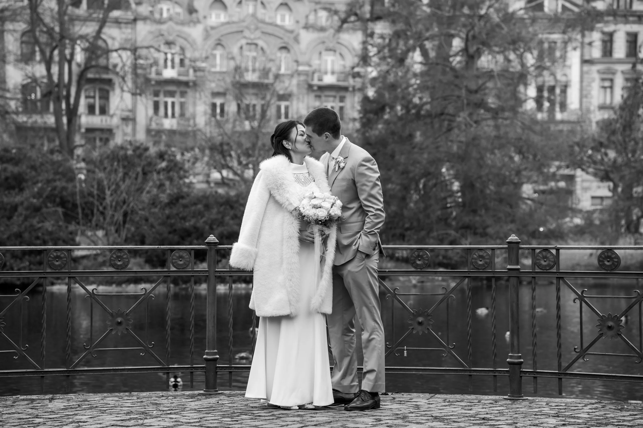 Braut und Bräutigam bei Hochzeitsaufnahme am Flussufer, sie küssen sich, Braut hält einen Blumenstrauß, im Hintergrund Bäume und historische Gebäude, schwarz-weiß Foto.