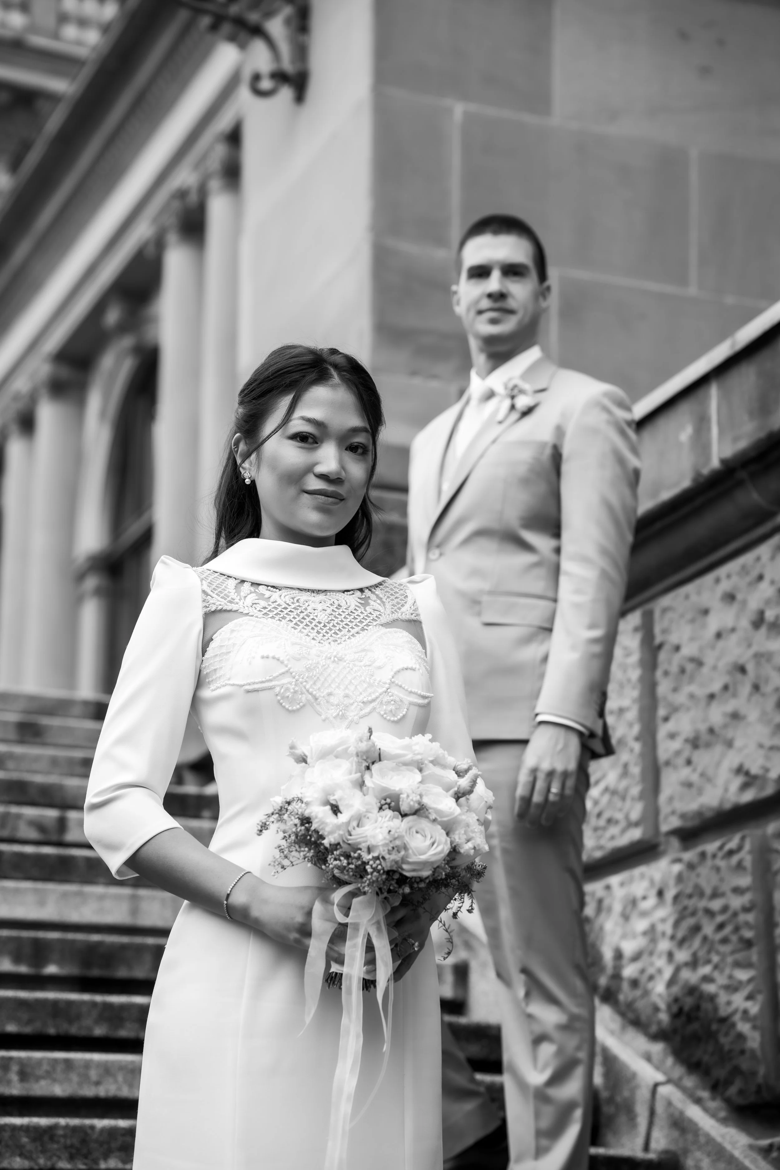 Ein Brautpaar steht auf Treppen vor einem großen gemauerten Gebäude. Die Braut hält einen Blumenstrauß und trägt ein weißes Hochzeitskleid, der Bräutigam trägt einen hellen Anzug.