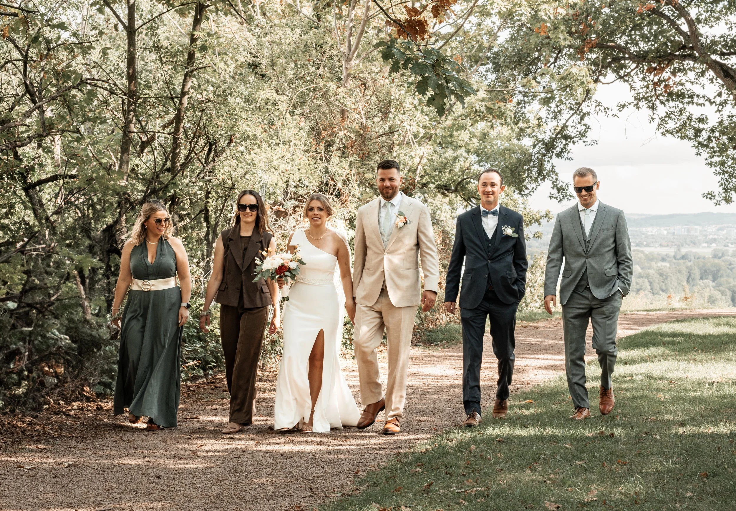 Hochzeitspaar und Gäste beim Spaziergang im Park bei Sonnenlicht, Bäume im Hintergrund.