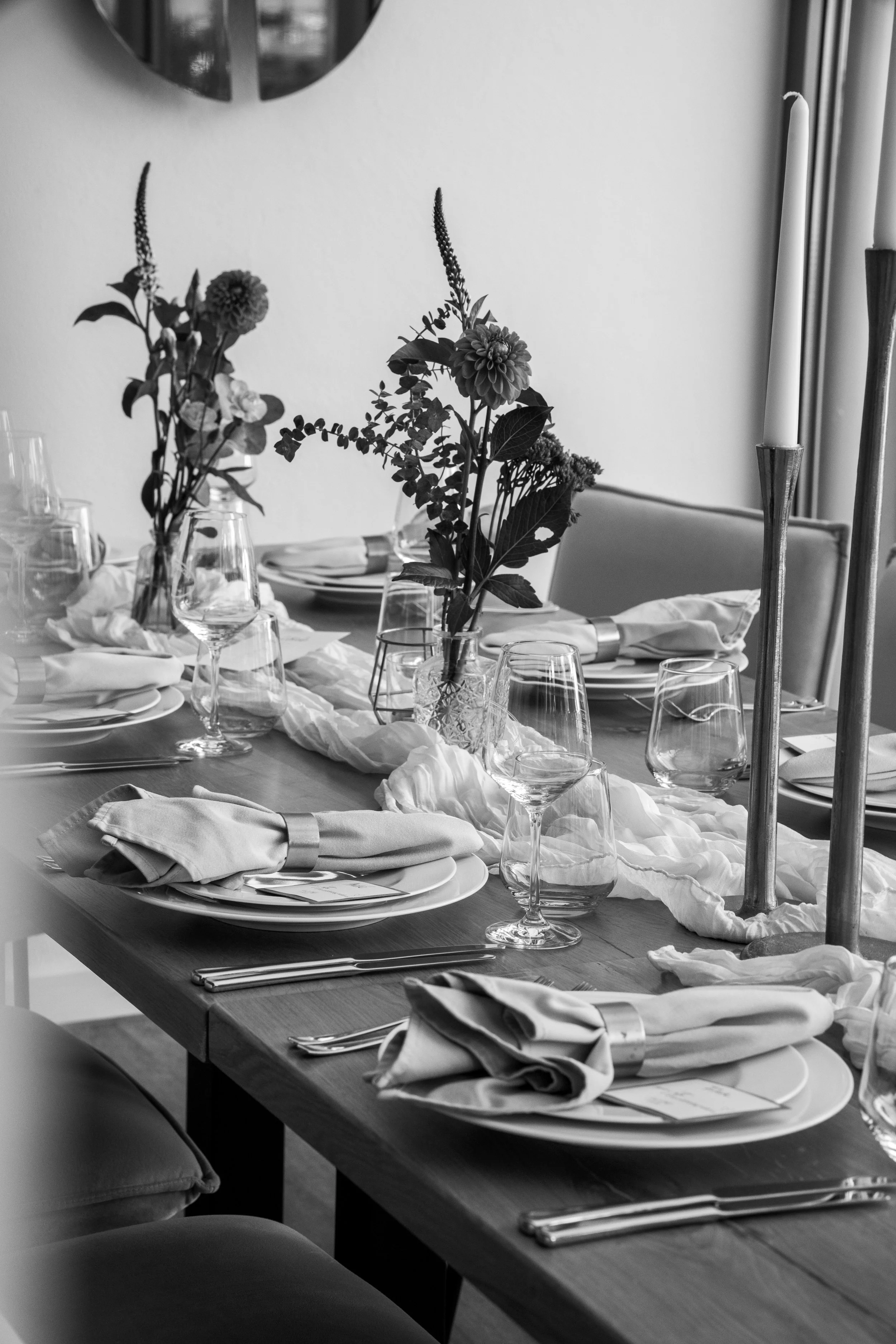 Ein gedeckter Esstisch mit Wein- und Wassergläsern, weißen Tellern und Besteck, verziert mit Blumen in Vasen und Kerzen, schwarz-weiße Farbgestaltung.